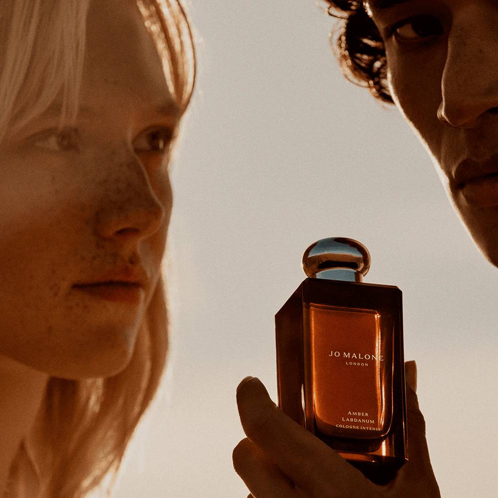 Um homem e uma mulher, com a pele iluminada por um p&ocirc;r do sol dourado, observam um frasco de perfume Jo Malone London Amber & Labdanum Cologne Intense, segurado pela m&atilde;o do homem. O frasco retangular tem l&iacute;quido &acirc;mbar e tampa esf&eacute;rica prateada.