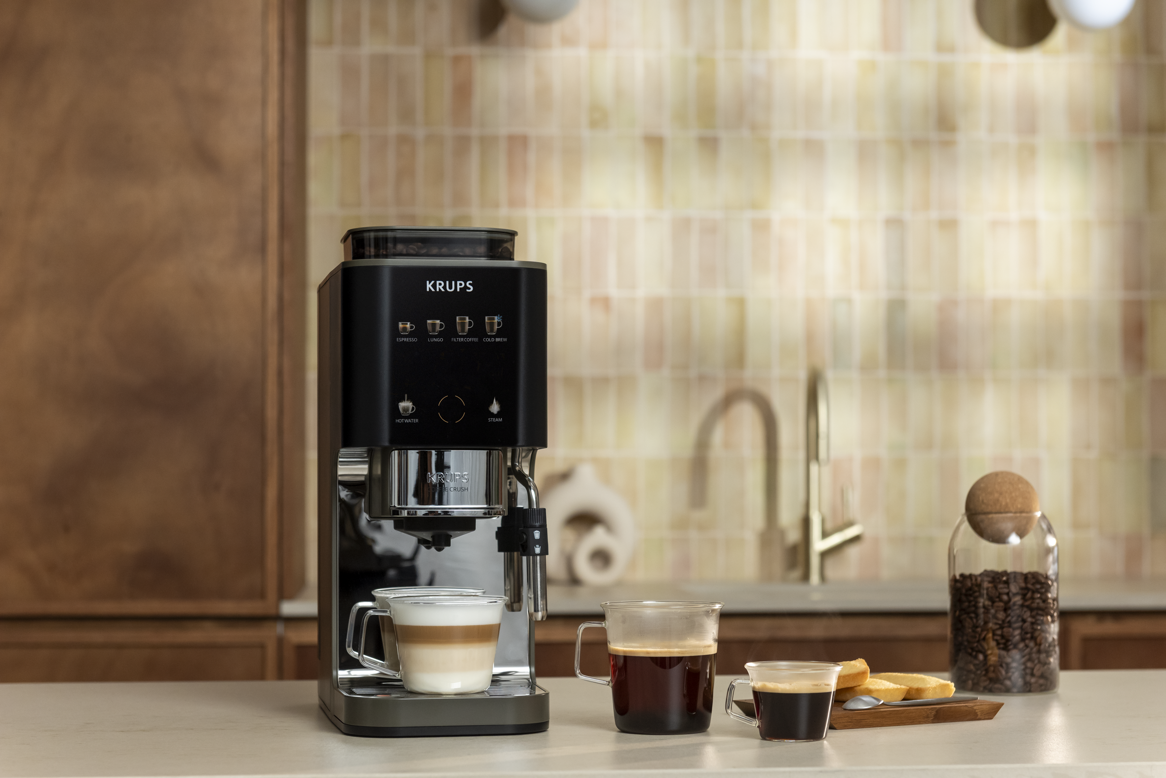 Máquina de café preta Krups em uma bancada clara, com um copo de café latte, dois copos de café preto, torradas e um pote de grãos de café. Ao fundo, uma cozinha com armários de madeira e azulejos bege