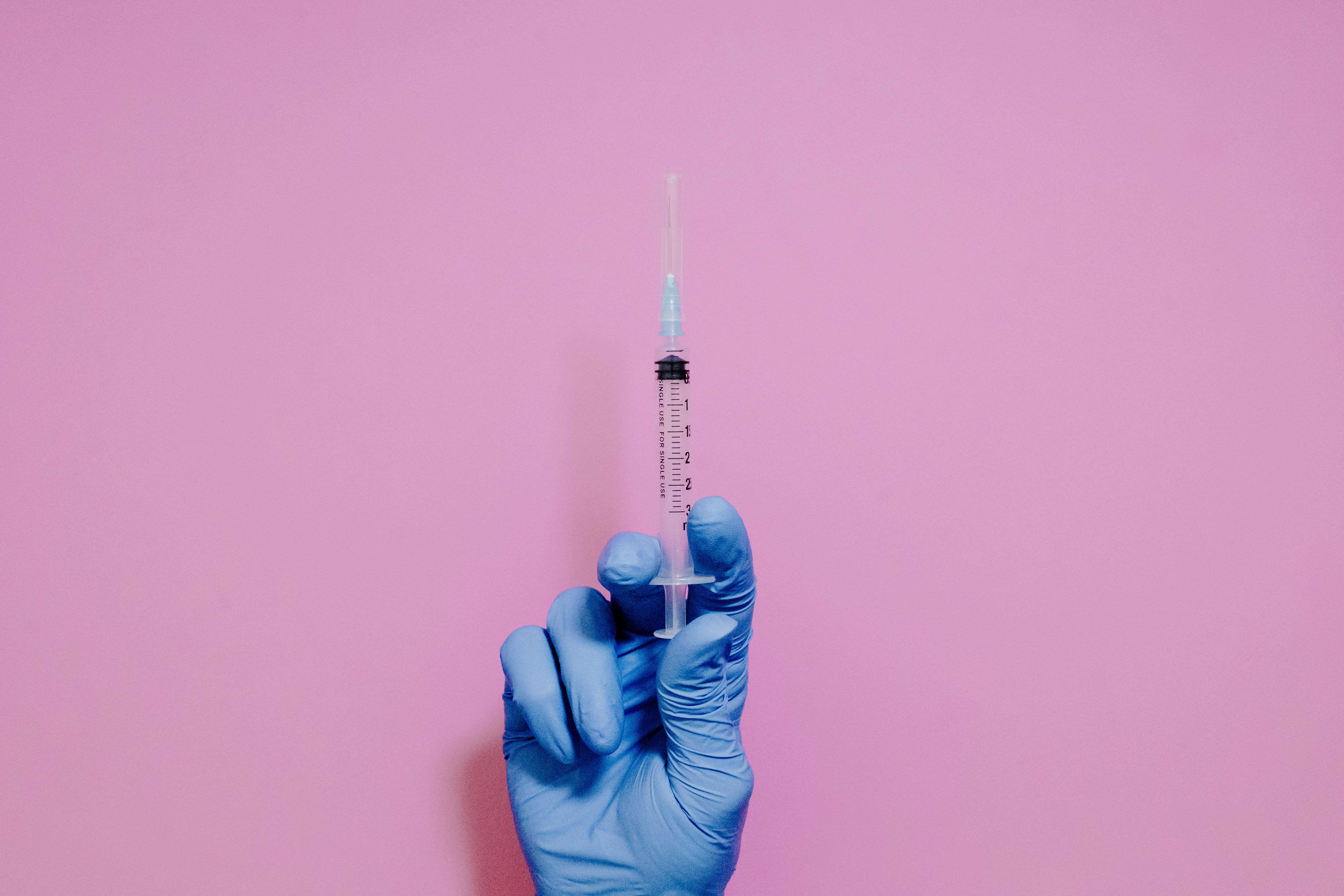 Mão enluvada de azul segura uma seringa transparente com agulha, contra um fundo rosa liso