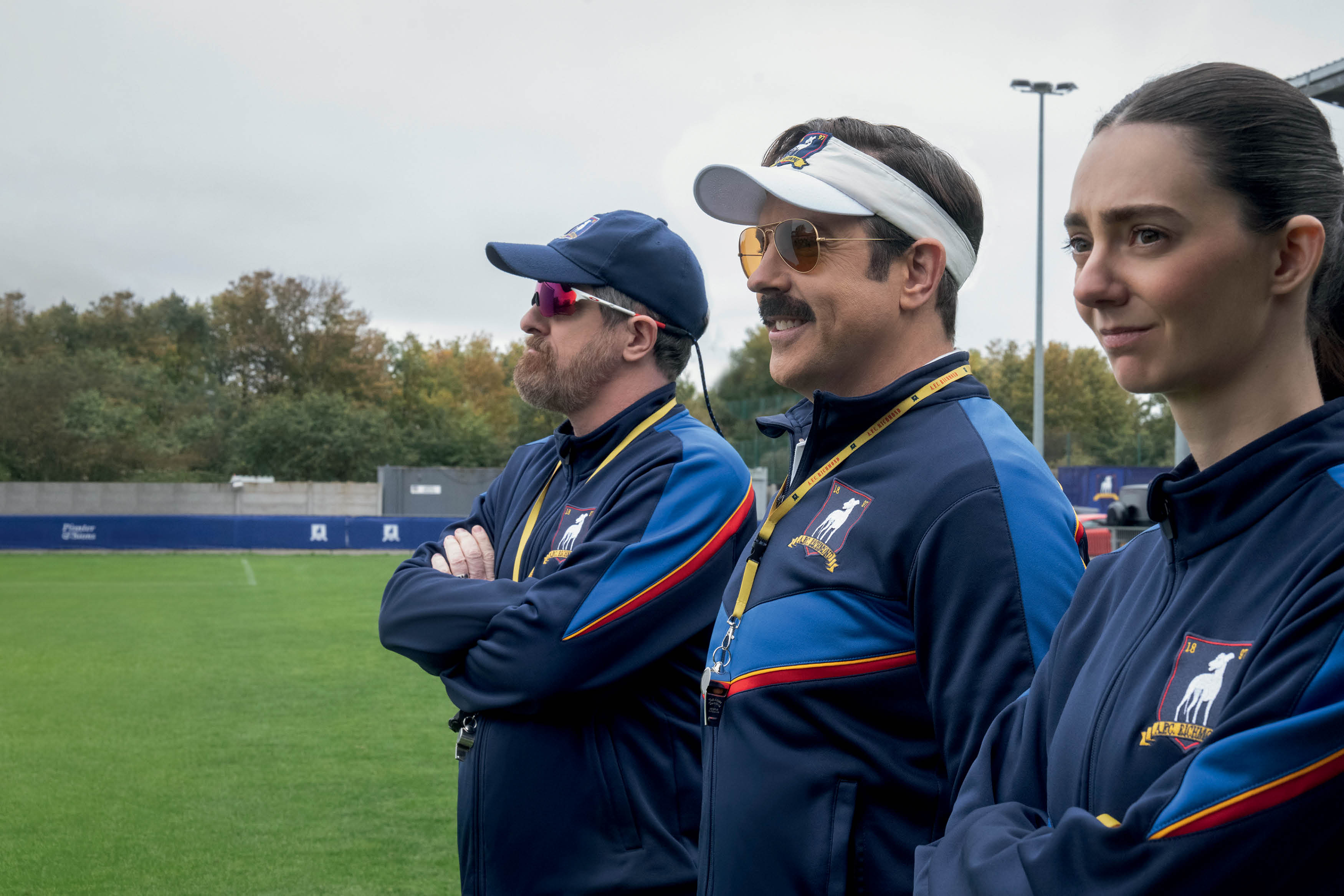 Três pessoas, dois homens e uma mulher, em uniformes esportivos azuis com listras vermelhas e azuis, estão em um campo de futebol gramado. O homem do meio usa um boné branco, óculos de sol e bigode, sorrindo levemente. A mulher à direita tem cabelo escuro preso e expressão séria. O homem à esquerda usa boné azul e óculos esportivos, com barba. Todos olham para a direita, com os braços cruzados, em um dia nublado