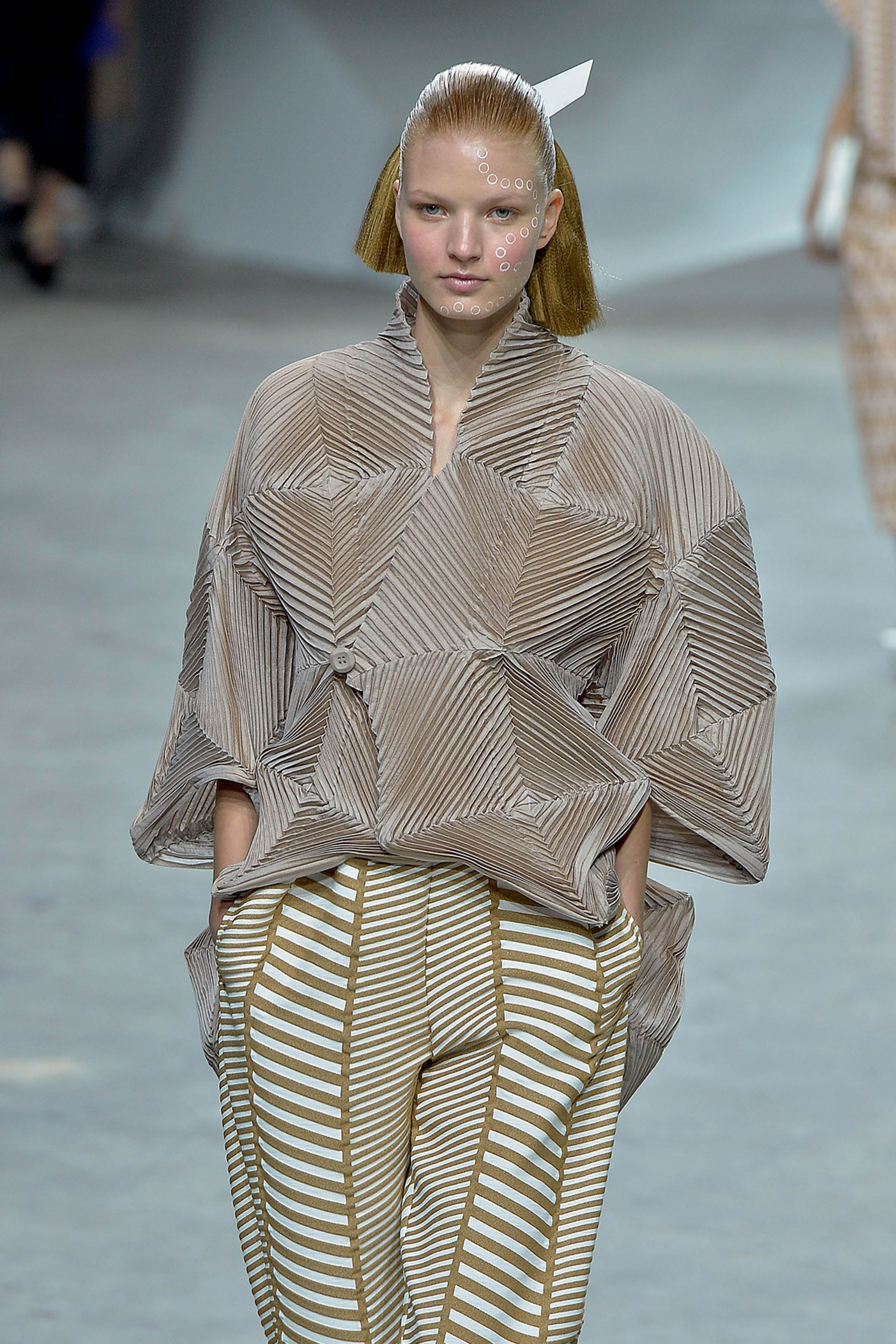 Modelo com cabelo loiro curto e liso, maquiagem de c&iacute;rculos brancos no rosto, vestindo blusa bege plissada com formas geom&eacute;tricas e cal&ccedil;a com listras brancas e marrons em zigue-zague, posando em passarela cinza