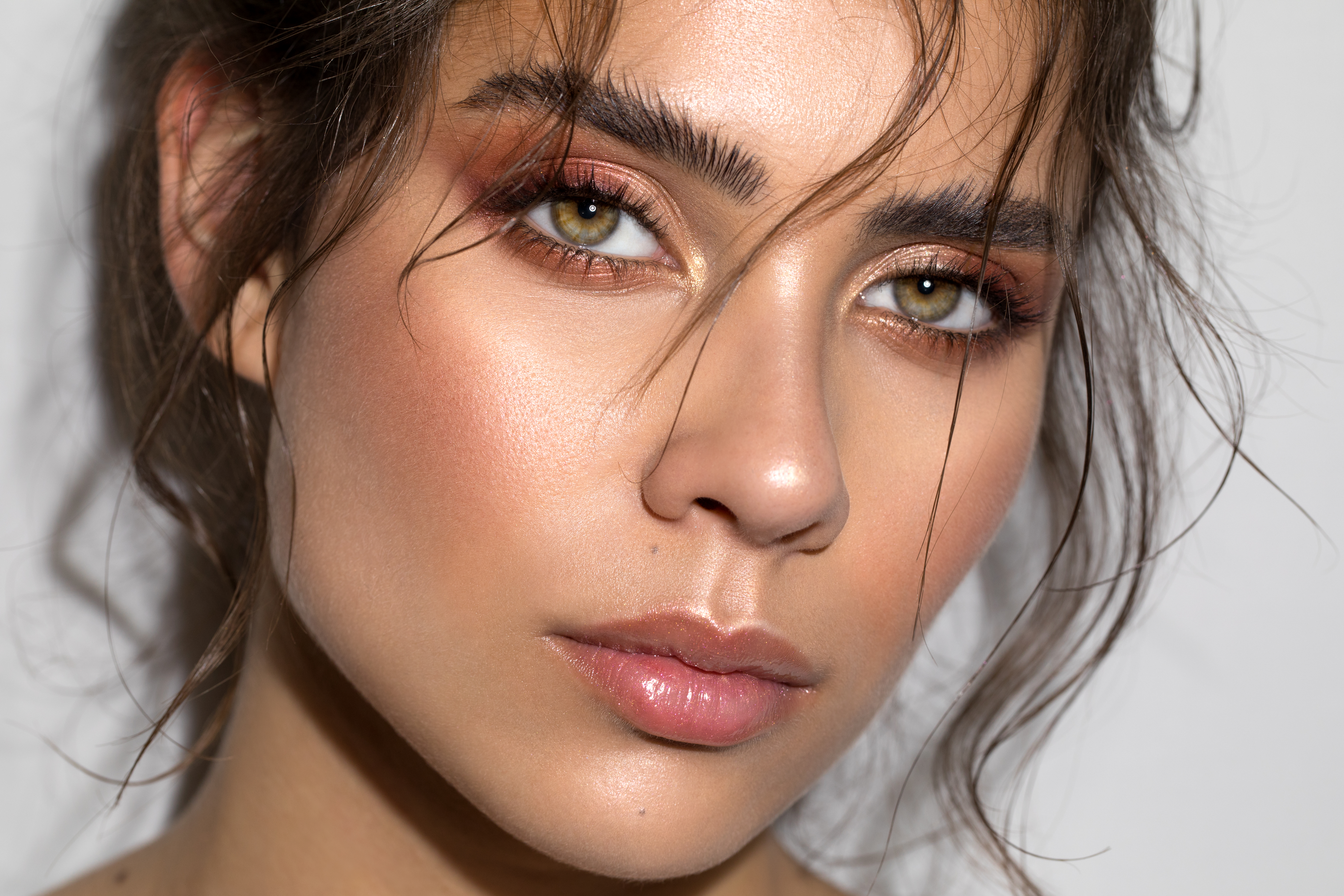 Rosto de uma mulher jovem com maquiagem em tons terrosos, olhos verdes e lábios rosados. Cabelos castanhos soltos emolduram o rosto, com alguns fios sobre a testa e bochechas. A pele tem um brilho natural, e as sobrancelhas são cheias.