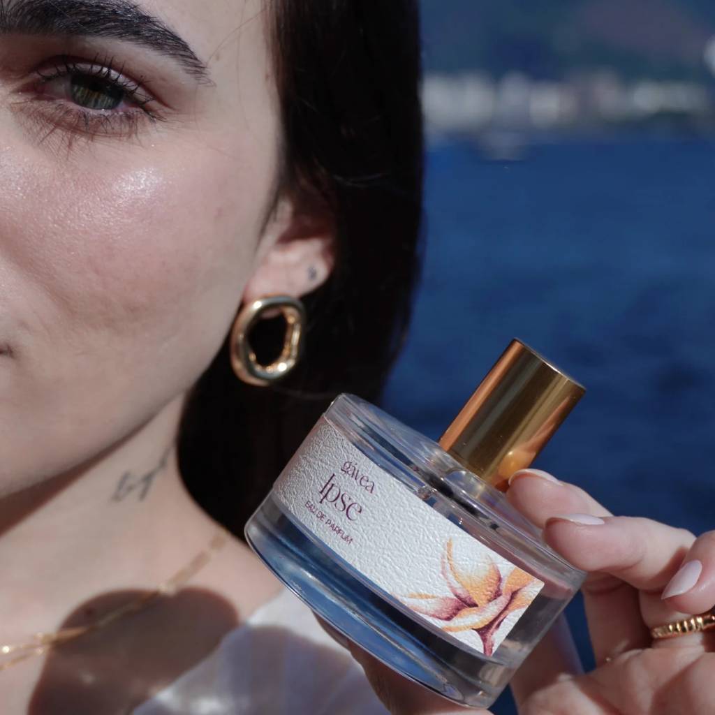 Close-up de uma mulher com cabelo escuro e brinco dourado, segurando um frasco de perfume G&aacute;vea Ipse Eau de Parfum com r&oacute;tulo floral, contra um fundo azul desfocado