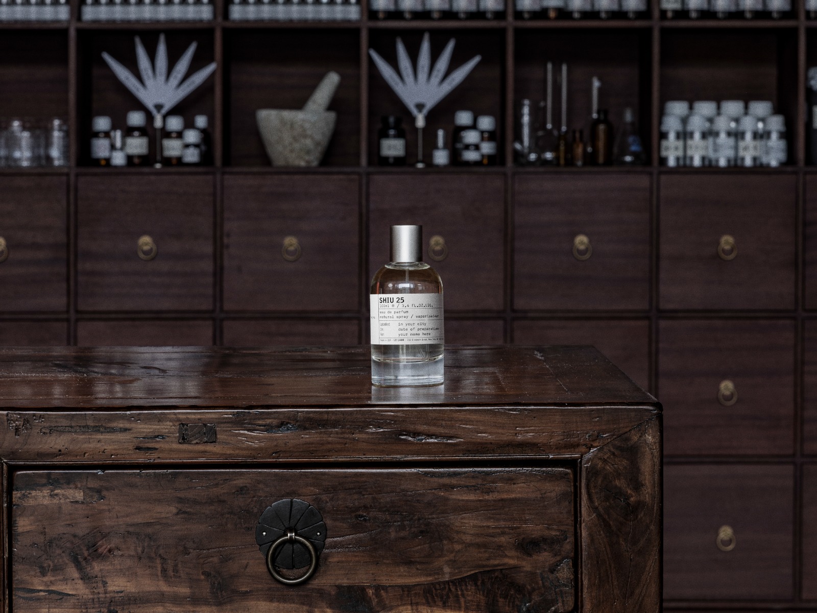 Frasco de perfume transparente com rótulo branco SHIU 25 sobre uma mesa de madeira escura e rústica, com um armário de gavetas e nichos com frascos e objetos de laboratório ao fundo