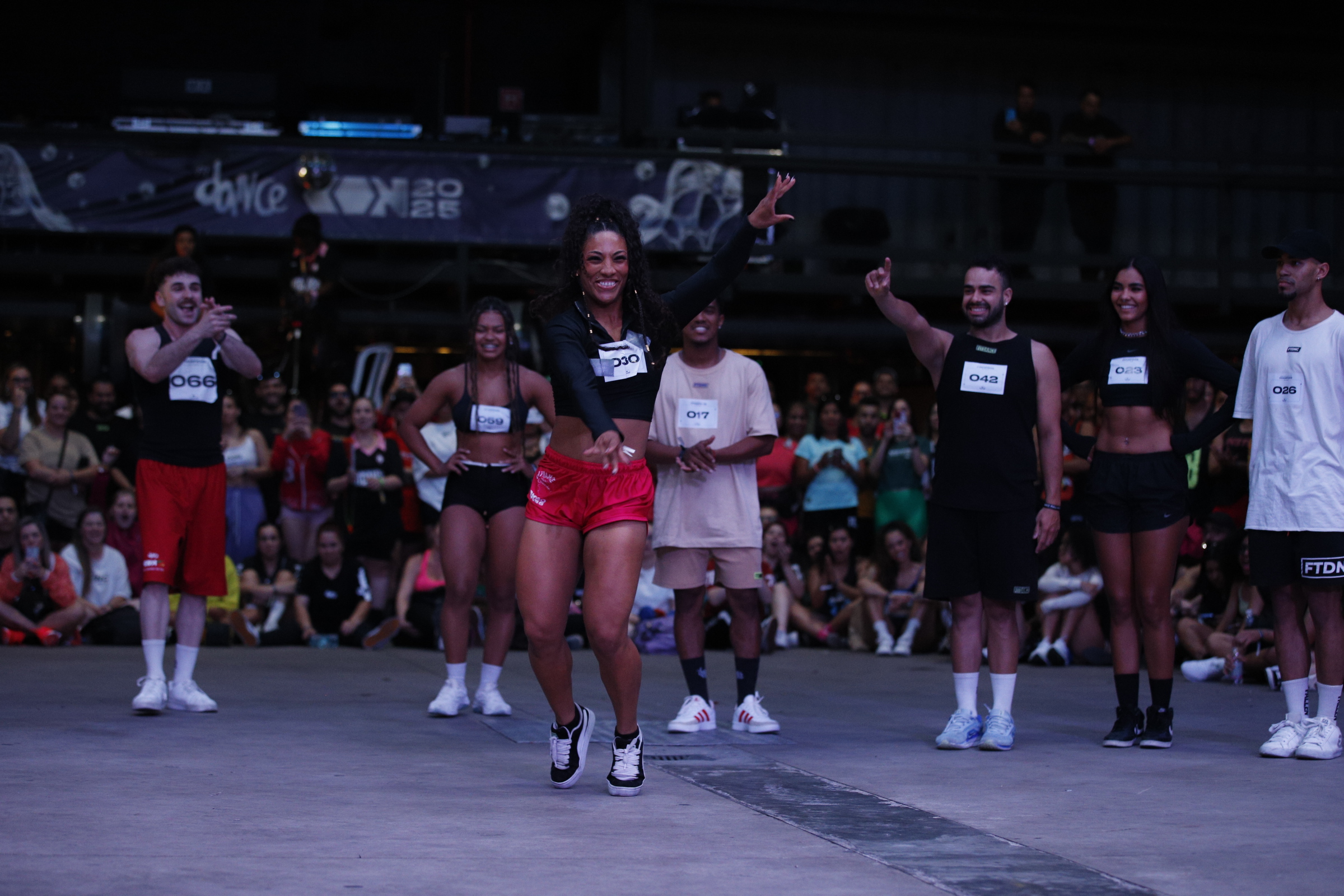 Mulher negra sorrindo, com cabelo cacheado preso, blusa preta e shorts vermelhos, dança no centro de um palco. Ao redor, outros dançarinos e uma plateia sentada no chão, com um banner Dance 2025 ao fundo