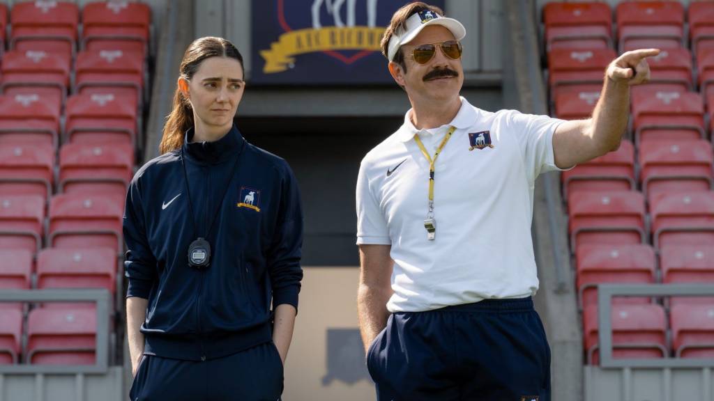 Um homem e uma mulher, ambos com uniformes esportivos, em um est&aacute;dio com cadeiras vermelhas. O homem, &agrave; direita, usa &oacute;culos de sol, bon&eacute; branco, camisa polo branca e apito, apontando para a direita com um sorriso. A mulher, &agrave; esquerda, usa agasalho azul-marinho e um cron&ocirc;metro no pesco&ccedil;o, com express&atilde;o s&eacute;ria