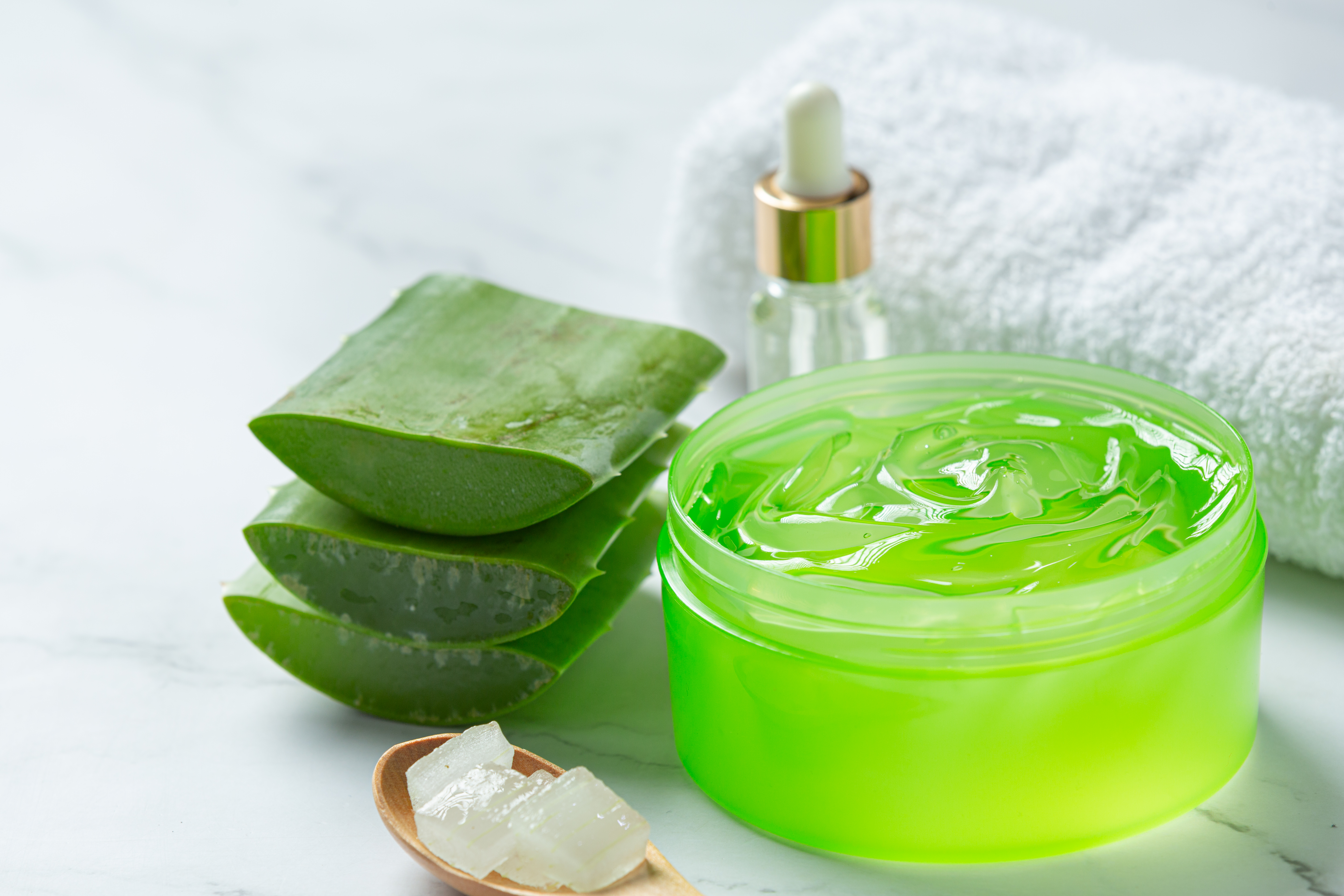 Folhas de aloe vera cortadas, um pote de gel verde, um frasco conta-gotas e uma toalha branca sobre uma superf&iacute;cie de m&aacute;rmore claro. Uma colher de madeira com peda&ccedil;os do gel da planta est&aacute; em primeiro plano