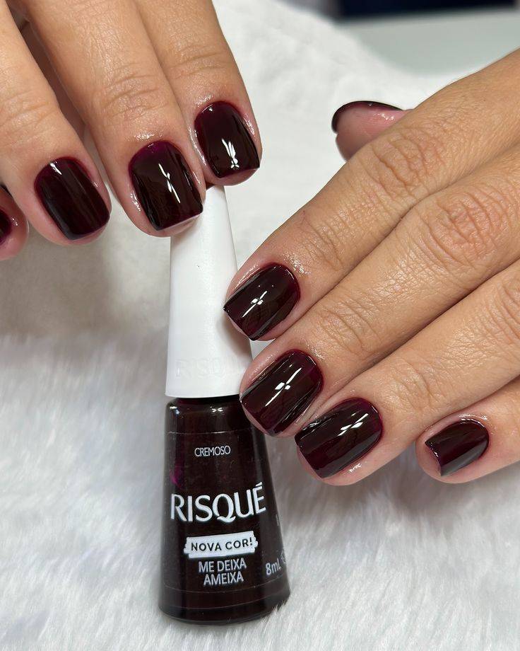 M&atilde;o feminina com unhas curtas pintadas de esmalte vinho escuro e brilhante, segurando um frasco de esmalte Risqu&eacute; Me Deixa Ameixa