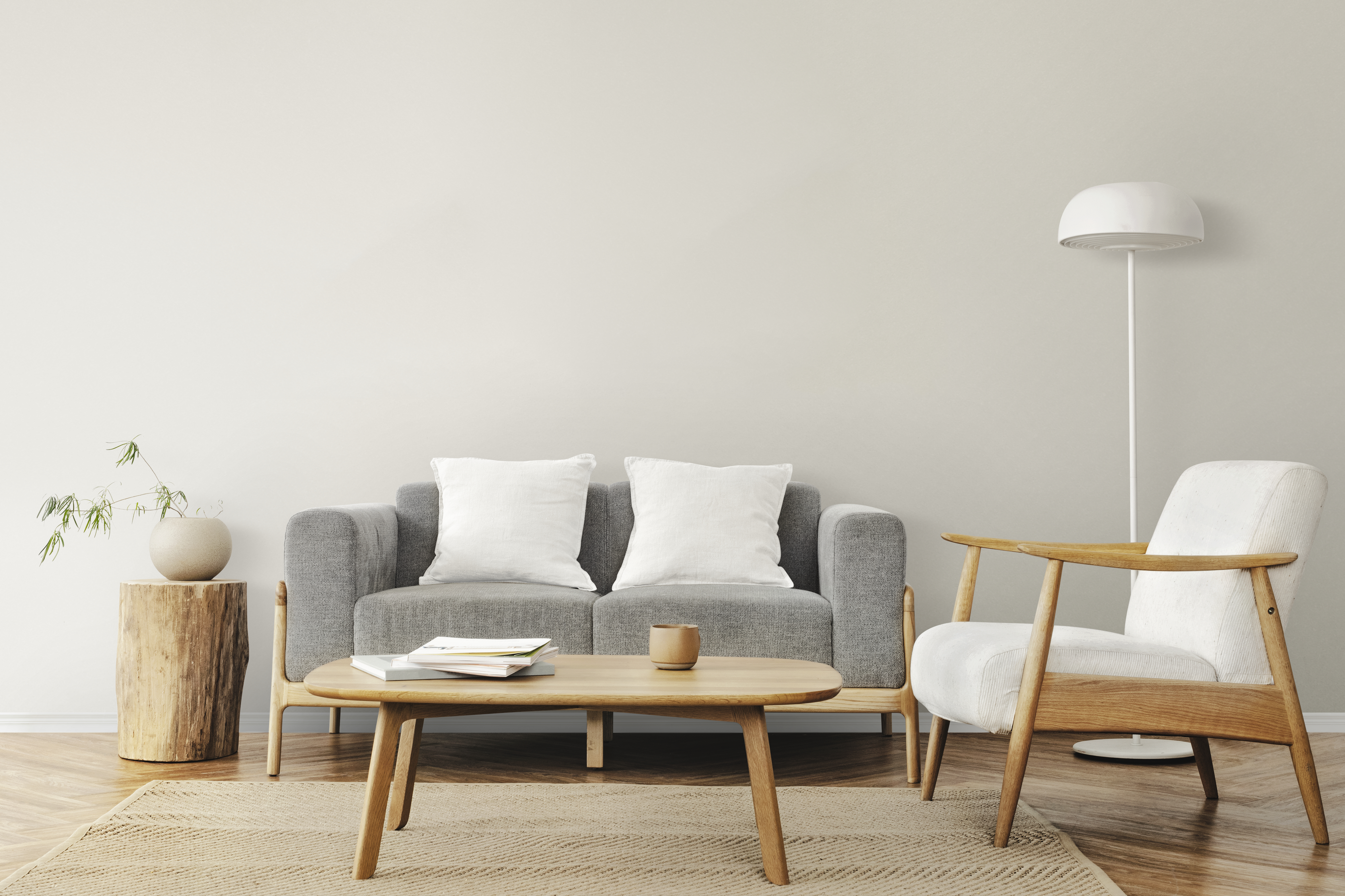 Sala de estar minimalista com sofá cinza de dois lugares e almofadas brancas, poltrona branca com estrutura de madeira clara, mesa de centro oval de madeira com livros e xícara, e luminária de chão branca. Um vaso com galhos verdes está sobre um tronco de madeira ao lado do sofá. O piso é de madeira e há um tapete bege com textura geométrica. A parede é lisa e clara