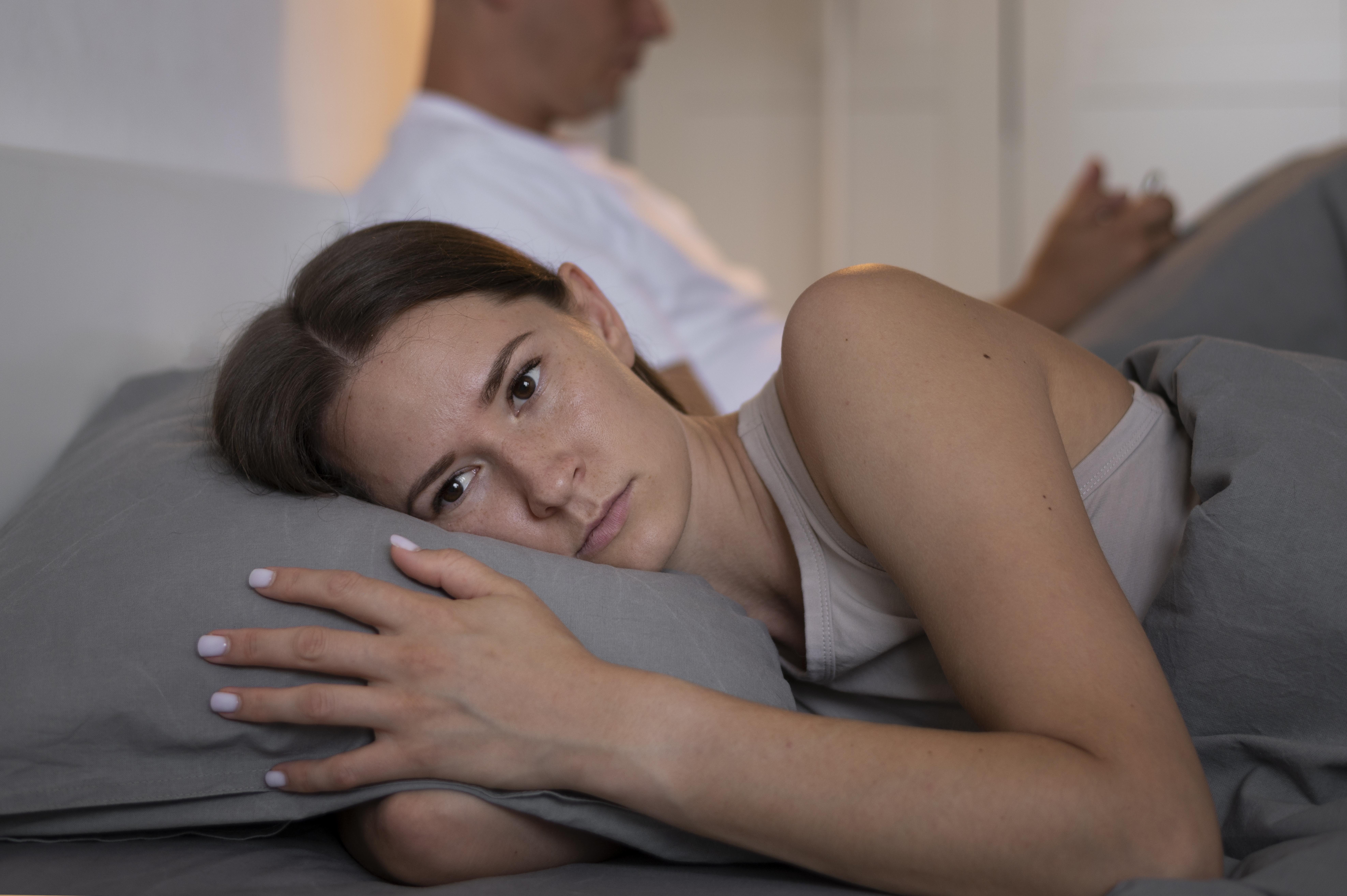 Mulher deitada na cama, com express&atilde;o triste, olhando para frente, enquanto um homem de costas para ela usa o celular ao fundo