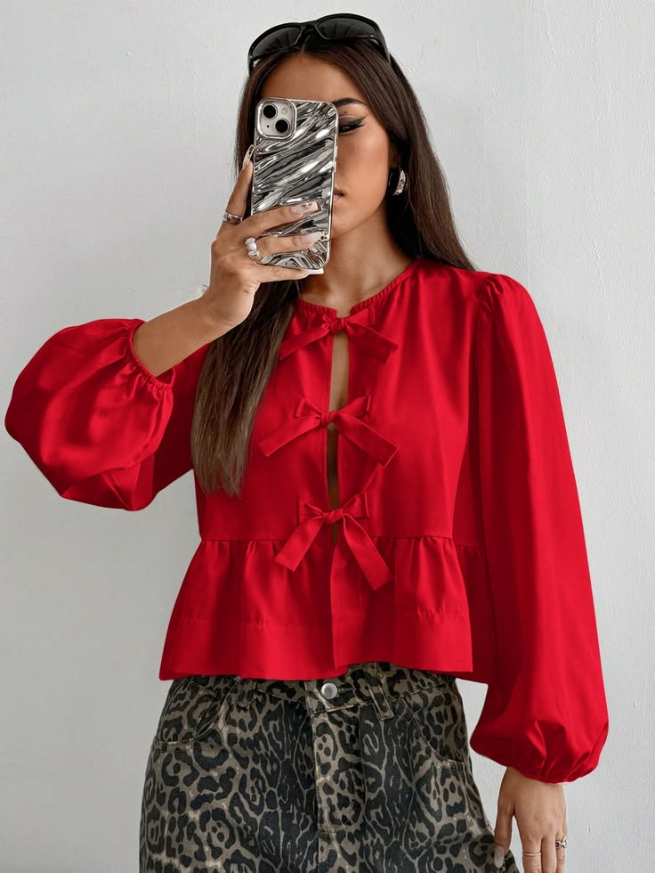Mulher com cabelo castanho escuro, &oacute;culos de sol na cabe&ccedil;a, usando blusa vermelha com la&ccedil;os e cal&ccedil;a estampada de oncinha, tirando uma selfie com o celular prateado que cobre parte do rosto