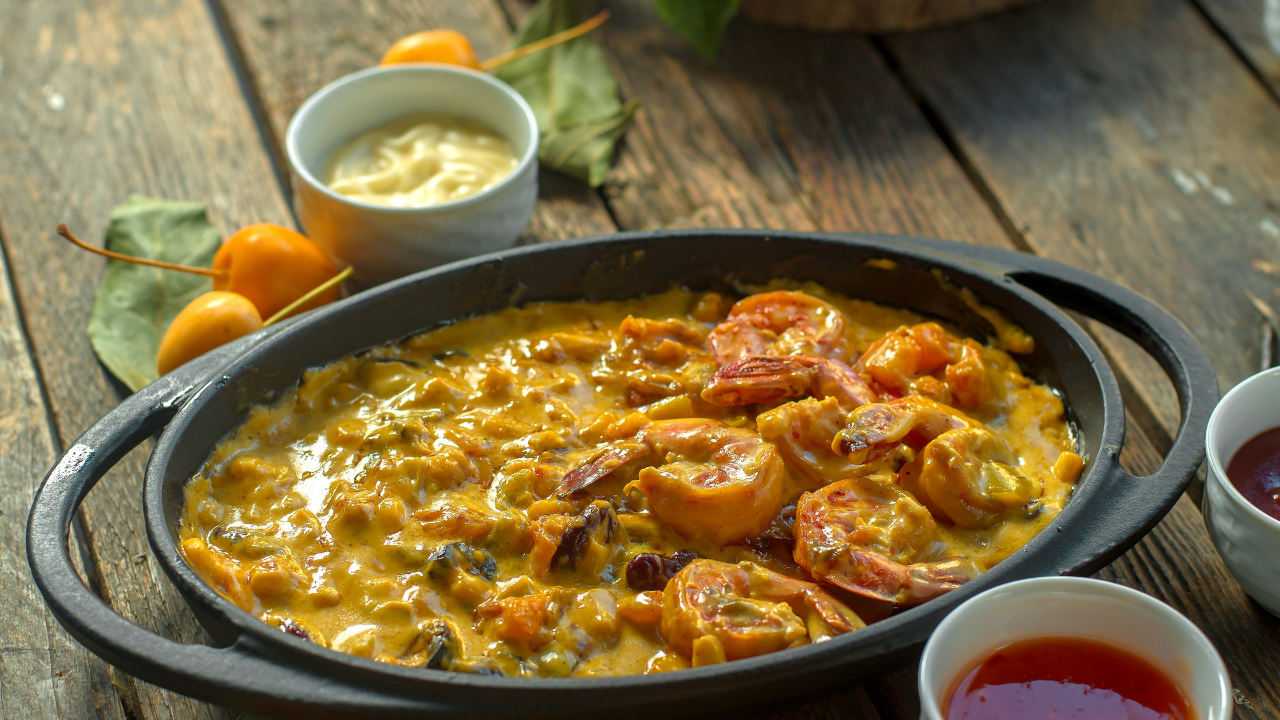 Moqueca de camarão em panela de ferro preta, com molho amarelo cremoso e pedaços de camarão, sobre mesa de madeira rústica. Ao fundo, pequenas frutas amarelas e folhas verdes. À direita, dois potes brancos com molhos, um vermelho e outro marrom. À esquerda, um pote branco com molho amarelo claro