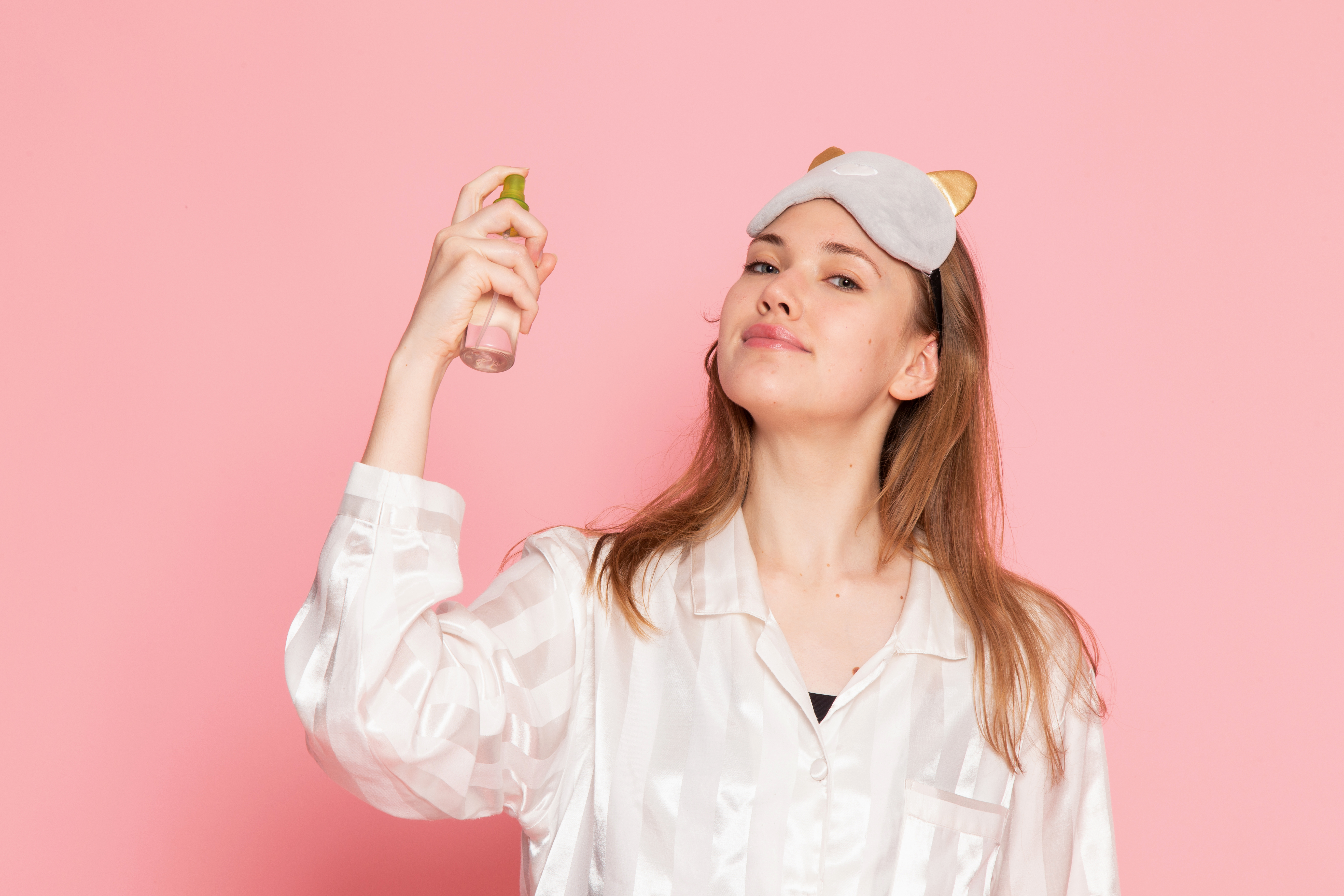 Mulher jovem de pijama branco e máscara de dormir cinza na cabeça, aplicando um spray facial com a mão direita, em fundo rosa