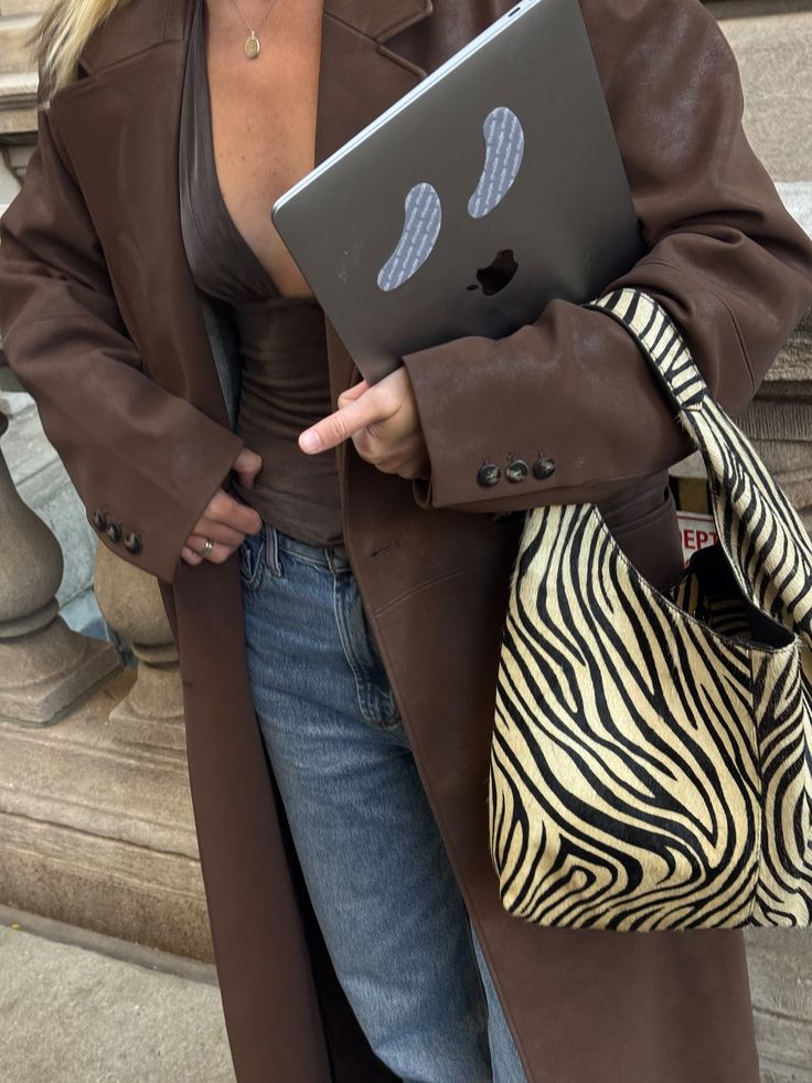 Pessoa com casaco marrom, blusa decotada, cal&ccedil;a jeans e bolsa de zebra, segurando um notebook cinza com adesivos de olhos de fantasma