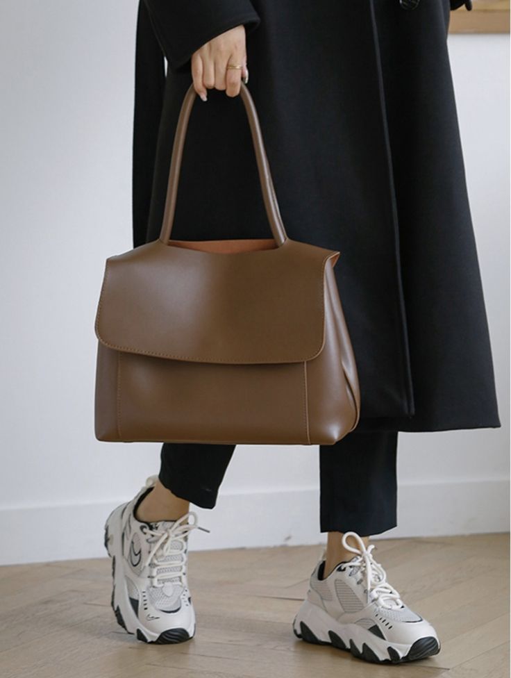 Pessoa com casaco preto e cal&ccedil;a escura, segurando uma bolsa marrom de couro sint&eacute;tico com a m&atilde;o direita, usando t&ecirc;nis brancos com detalhes pretos e solado robusto, em piso de madeira