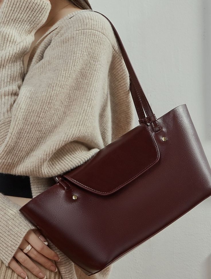 Mulher com su&eacute;ter bege de tric&ocirc;, usando uma bolsa de couro marrom-avermelhado com al&ccedil;a no ombro e fecho de aba, anel dourado no dedo anelar
