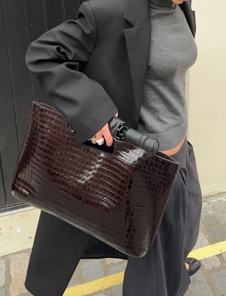 Pessoa com blazer preto, blusa cinza de gola alta e cal&ccedil;a escura, segurando uma bolsa marrom de couro com textura de crocodilo e um guarda-chuva preto dobrado. Usa an&eacute;is prateados e sapatilhas pretas, caminhando sobre cal&ccedil;amento de pedras com uma faixa amarela.