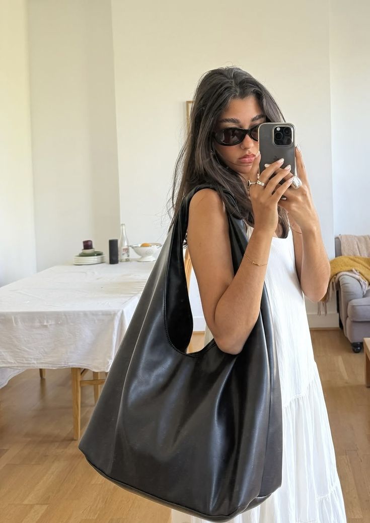 Mulher jovem de cabelos escuros e &oacute;culos de sol pretos, vestindo um vestido branco e carregando uma bolsa grande preta no ombro, tirando uma selfie com o celular em um ambiente interno claro