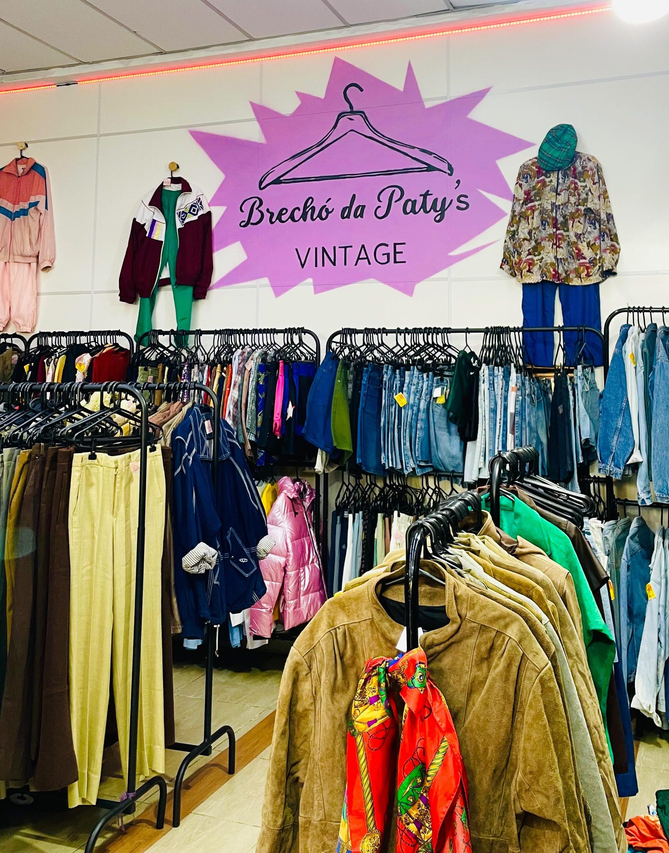 Interior de um brech&oacute; com araras cheias de roupas coloridas e vintage. Na parede branca, um letreiro roxo em formato de explos&atilde;o com um cabide desenhado e os dizeres Brech&oacute; da Patys VINTAGE. Tr&ecirc;s manequins exibem conjuntos de agasalho rosa e azul, jaqueta vinho com cal&ccedil;a verde, e camisa estampada com cal&ccedil;a azul e boina verde. O ch&atilde;o &eacute; de cer&acirc;mica clara e o teto tem lumin&aacute;rias fluorescentes