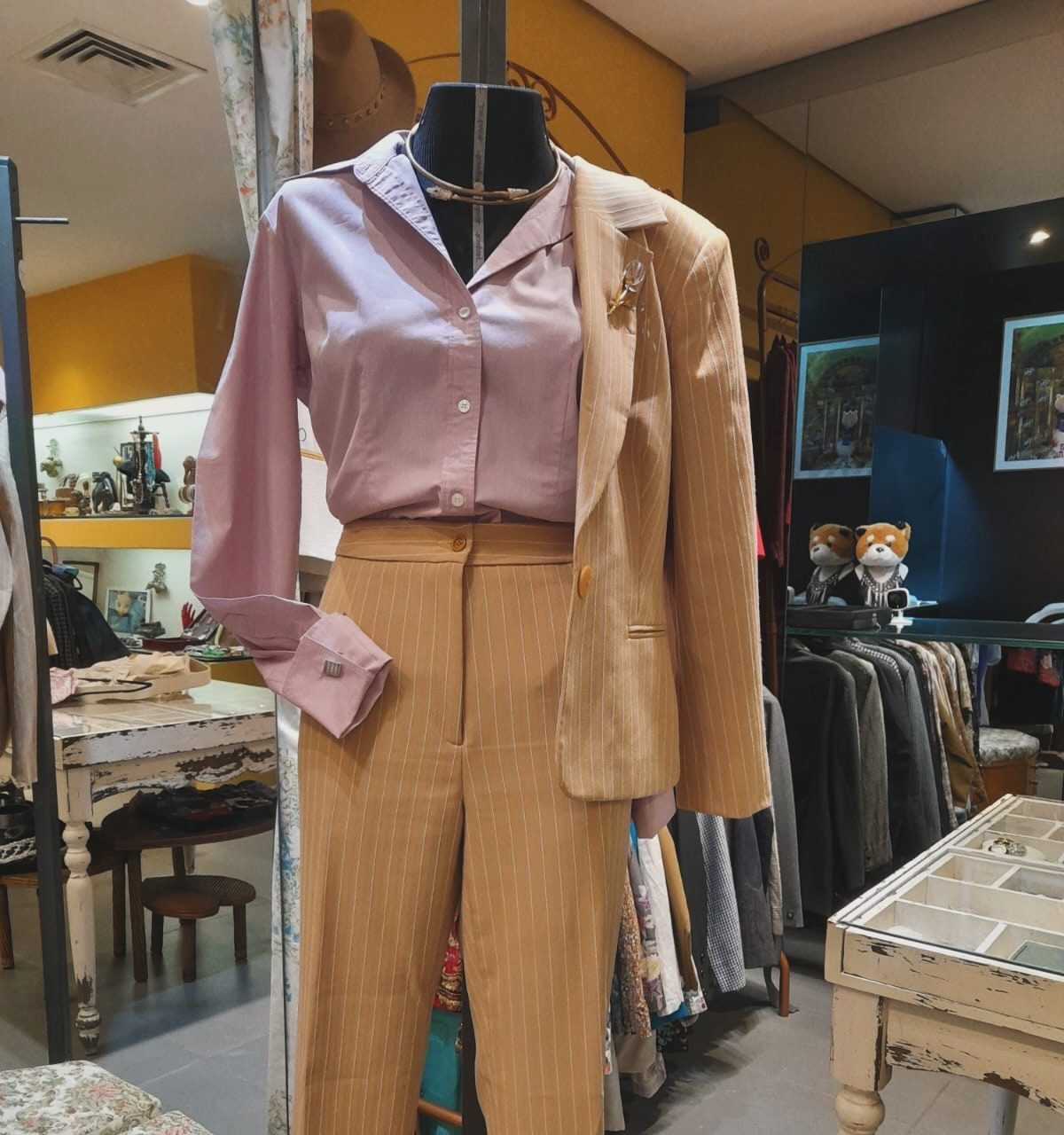 Manequim exibindo um conjunto de alfaiataria: camisa rosa-claro de manga longa com abotoaduras e cal&ccedil;a pantalona bege de risca de giz, com um blazer bege de risca de giz sobre o ombro direito, adornado com um broche dourado. Ao fundo, um ambiente de loja com roupas em araras e m&oacute;veis r&uacute;sticos