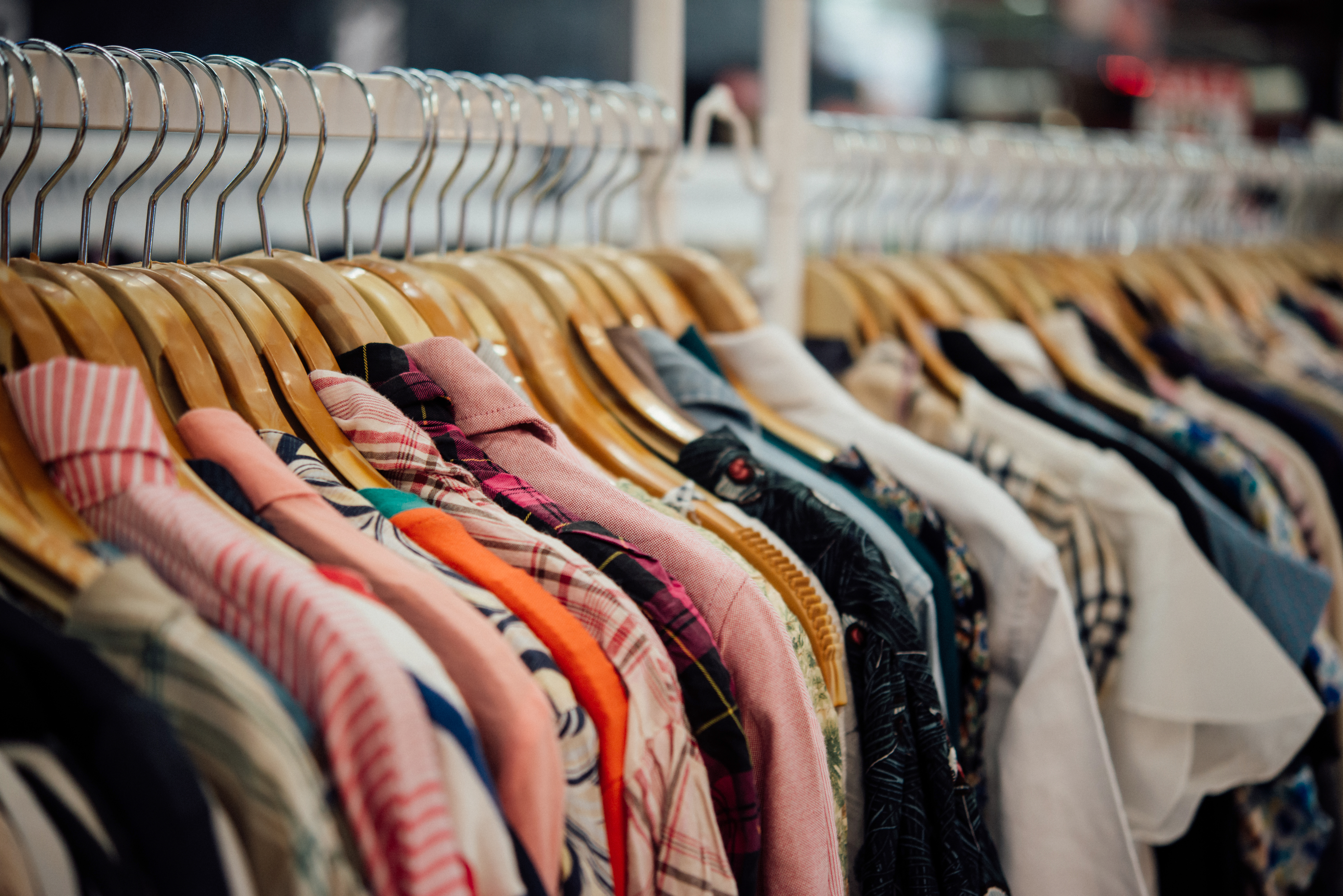 Uma fileira de camisas coloridas e estampadas penduradas em cabides de madeira e metal, em uma loja de roupas