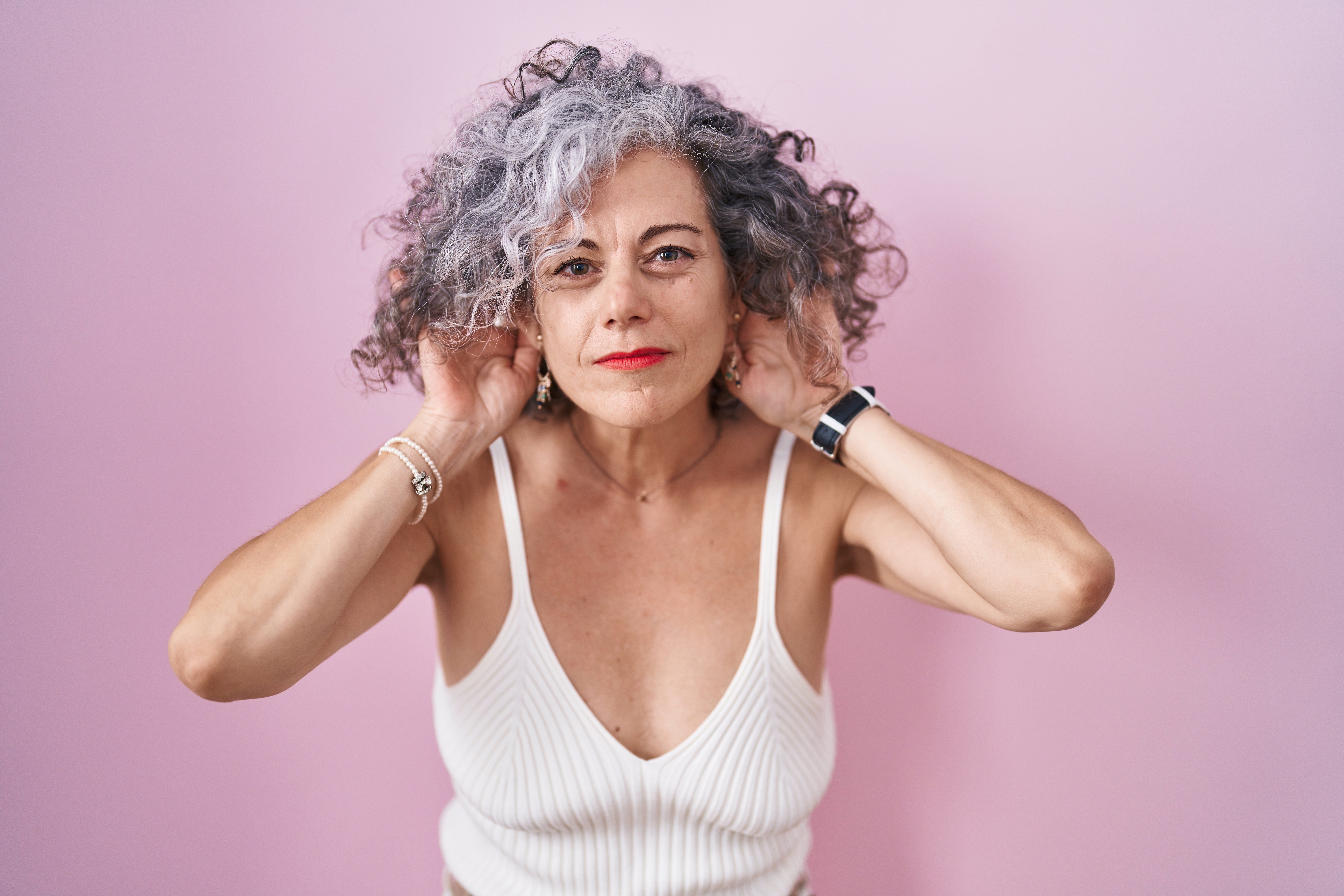 Mulher de meia-idade com cabelos grisalhos cacheados, blusa branca de alças e batom vermelho, segurando as orelhas com as mãos, em fundo rosa