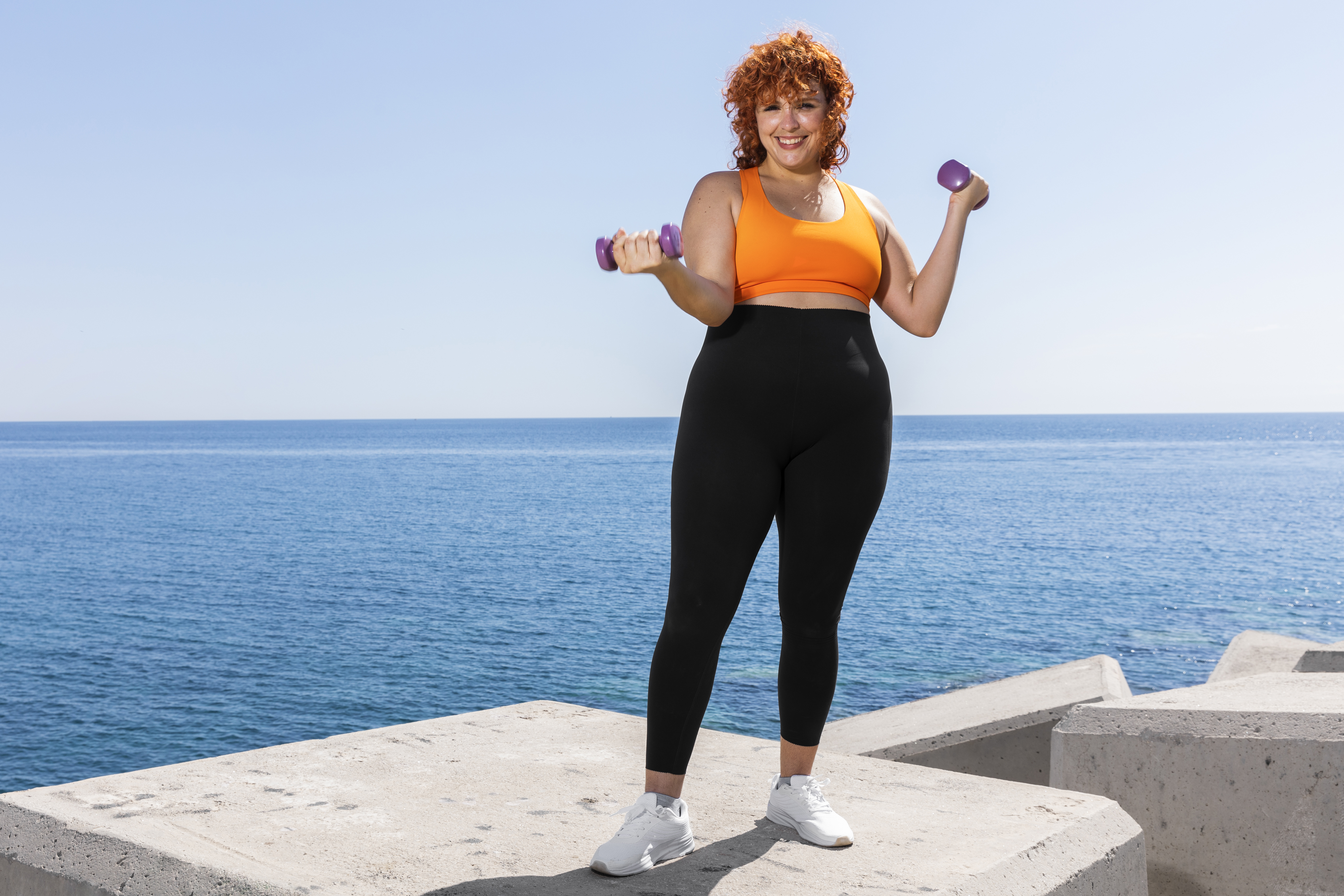 Mulher sorridente de cabelos ruivos cacheados, vestindo top laranja e calça legging preta, segurando halteres roxos. Ela está em um quebra-mar de concreto, com o mar azul e o céu claro ao fundo