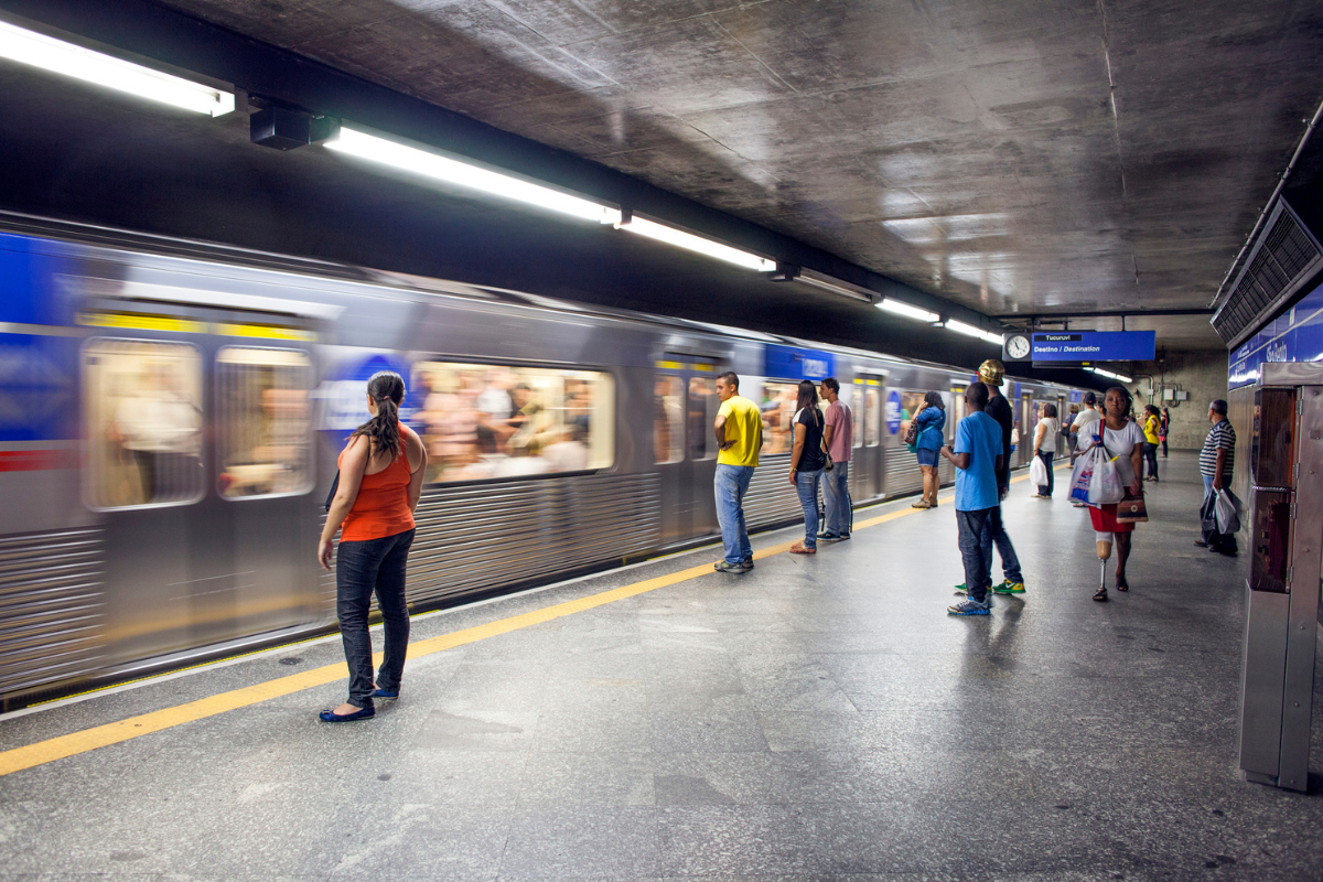 Pessoas aguardam um trem em movimento na plataforma de uma estação de metrô. Uma mulher de blusa laranja e calça escura está de costas, observando o trem. Outros passageiros, incluindo uma mulher com prótese na perna, estão espalhados pela plataforma, sob iluminação fluorescente. O trem prateado com faixa azul está em alta velocidade, com passageiros visíveis pelas janelas borradas. O teto de concreto aparente e a linha amarela de segurança na plataforma completam a cena