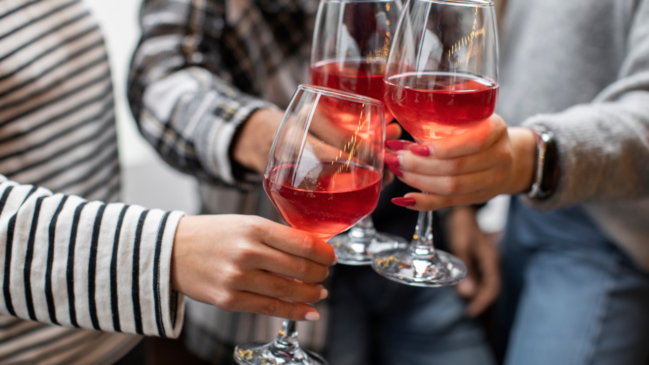 Três pessoas brindando com taças de vinho rosé, com as mãos visíveis. Uma pessoa veste blusa listrada preta e branca, outra camisa xadrez, e a terceira um suéter cinza com jeans. As unhas de duas pessoas estão pintadas de vermelho e rosa claro
