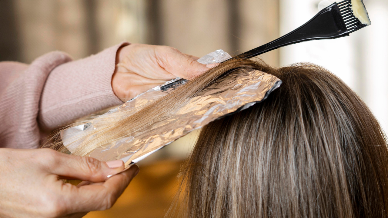 Uma pessoa com blusa rosa clara segura uma mecha de cabelo loiro em papel alumínio, enquanto outra mão aplica tintura com um pincel preto no cabelo castanho escuro da cliente, que tem alguns fios grisalhos visíveis