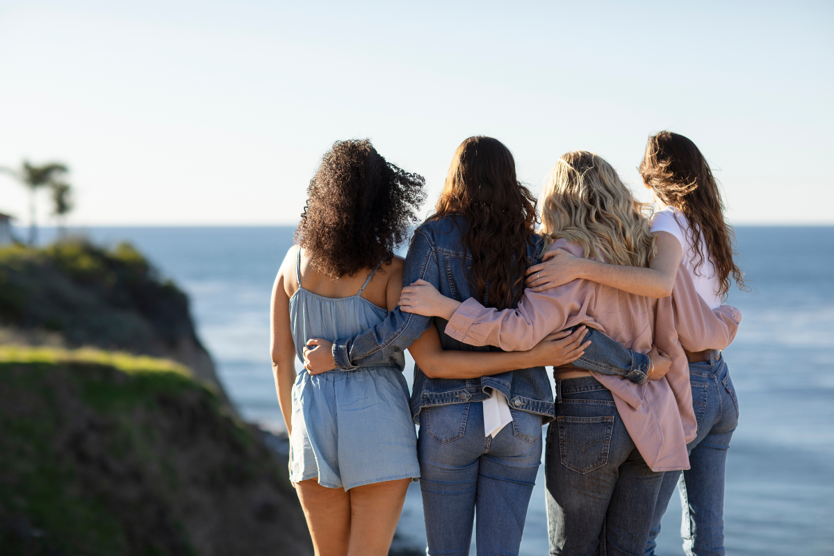 Quatro mulheres de costas, abraçadas, olhando o mar azul sob um céu claro. A primeira tem cabelo cacheado escuro e veste um macacão jeans claro. A segunda usa jaqueta e calça jeans. A terceira, com cabelo loiro ondulado, veste blusa rosa e calça jeans escura. A última, com cabelo castanho, usa camiseta branca e calça jeans