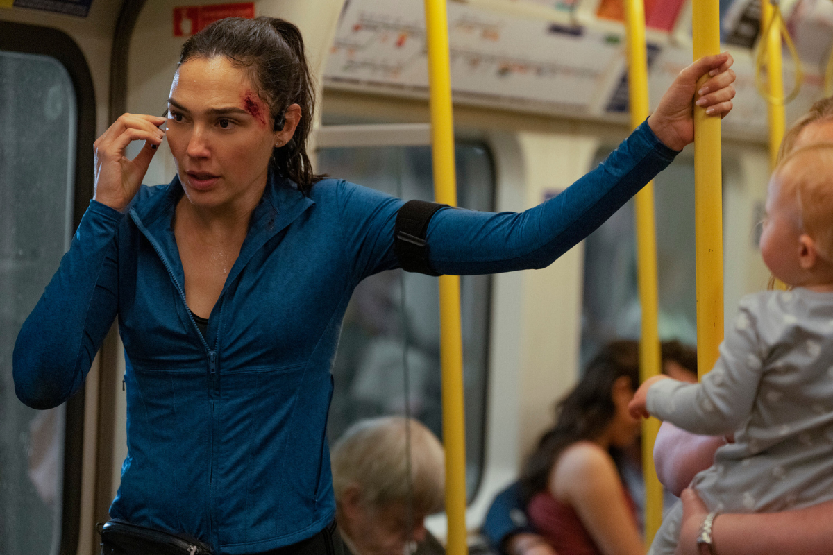 Mulher com cabelo preso, blusa esportiva azul e ferimento na testa, fala ao fone de ouvido enquanto segura uma barra amarela no metrô, olhando fixamente para frente. Um bebê está à direita, no colo de um adulto
