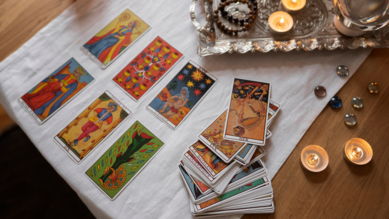 Cartas de tarô coloridas espalhadas sobre um pano branco, com um baralho empilhado à direita. Velas acesas e pedras decorativas em uma bandeja prateada e sobre a mesa de madeira complementam a cena