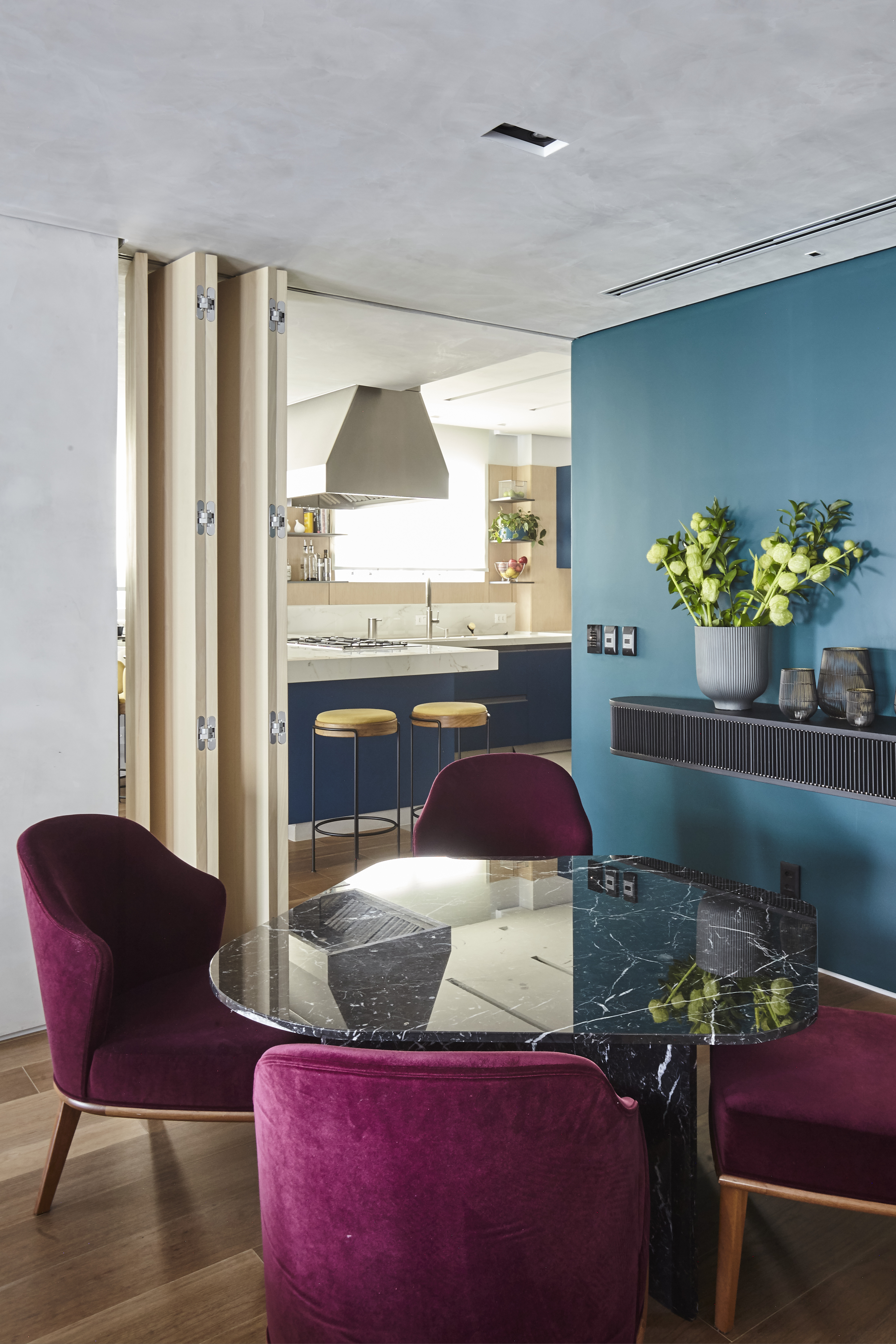 Sala de jantar com mesa redonda de m&aacute;rmore preto e quatro cadeiras estofadas em veludo vinho. Ao fundo, uma cozinha moderna com balc&atilde;o azul e banquetas amarelas, separada por uma porta sanfonada de madeira clara. Na parede azul-petr&oacute;leo, uma prateleira preta com um vaso de plantas verdes e objetos decorativos. O piso &eacute; de madeira clara e o teto cinza