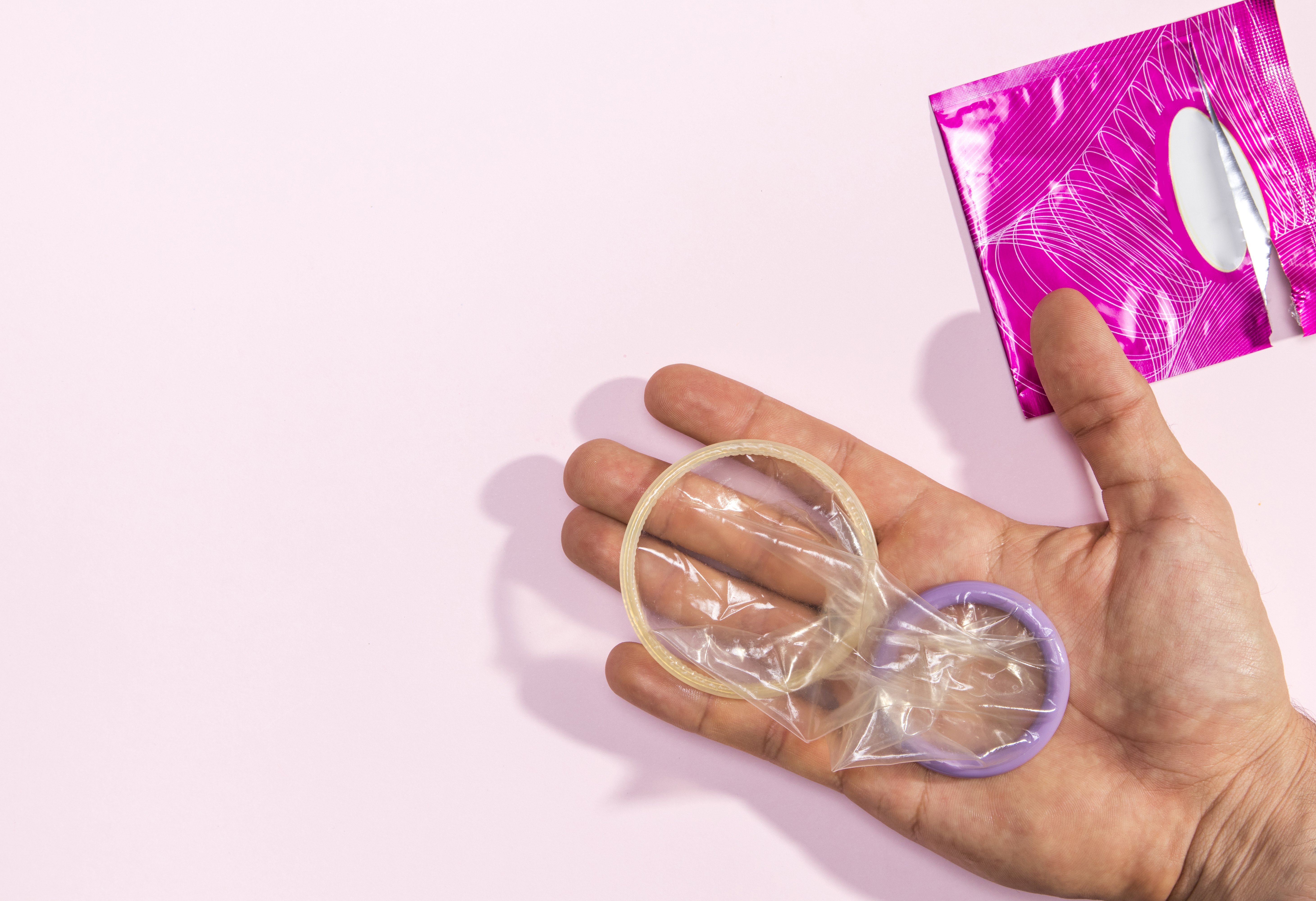 M&atilde;o masculina aberta segurando duas camisinhas abertas, uma com anel bege e outra com anel roxo, sobre fundo rosa claro. No canto superior direito, uma embalagem de camisinha rosa parcialmente aberta