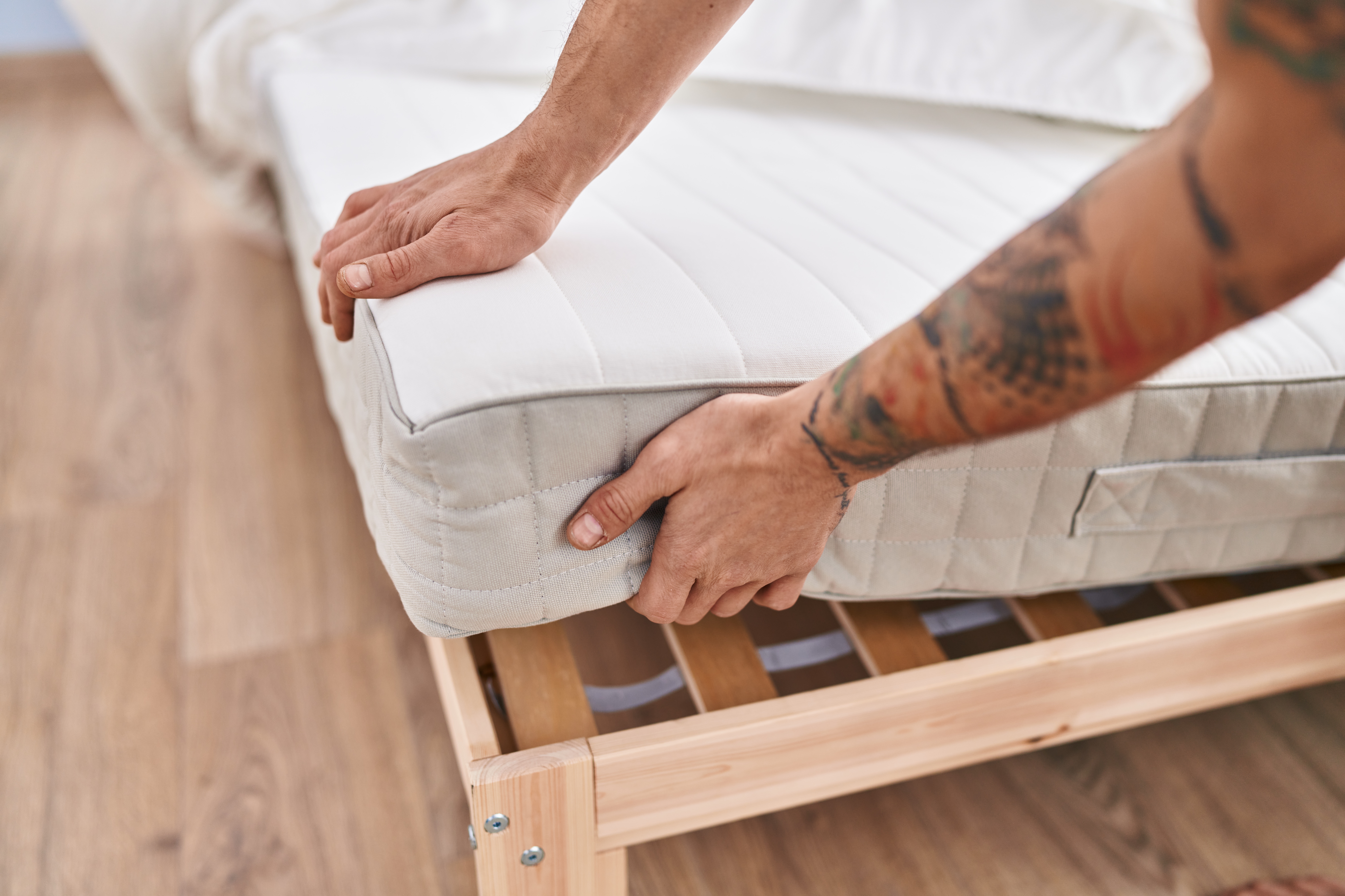 M&atilde;os de um homem com tatuagens no bra&ccedil;o direito ajustando um colch&atilde;o branco sobre uma estrutura de cama de madeira clara em um quarto com piso de madeira