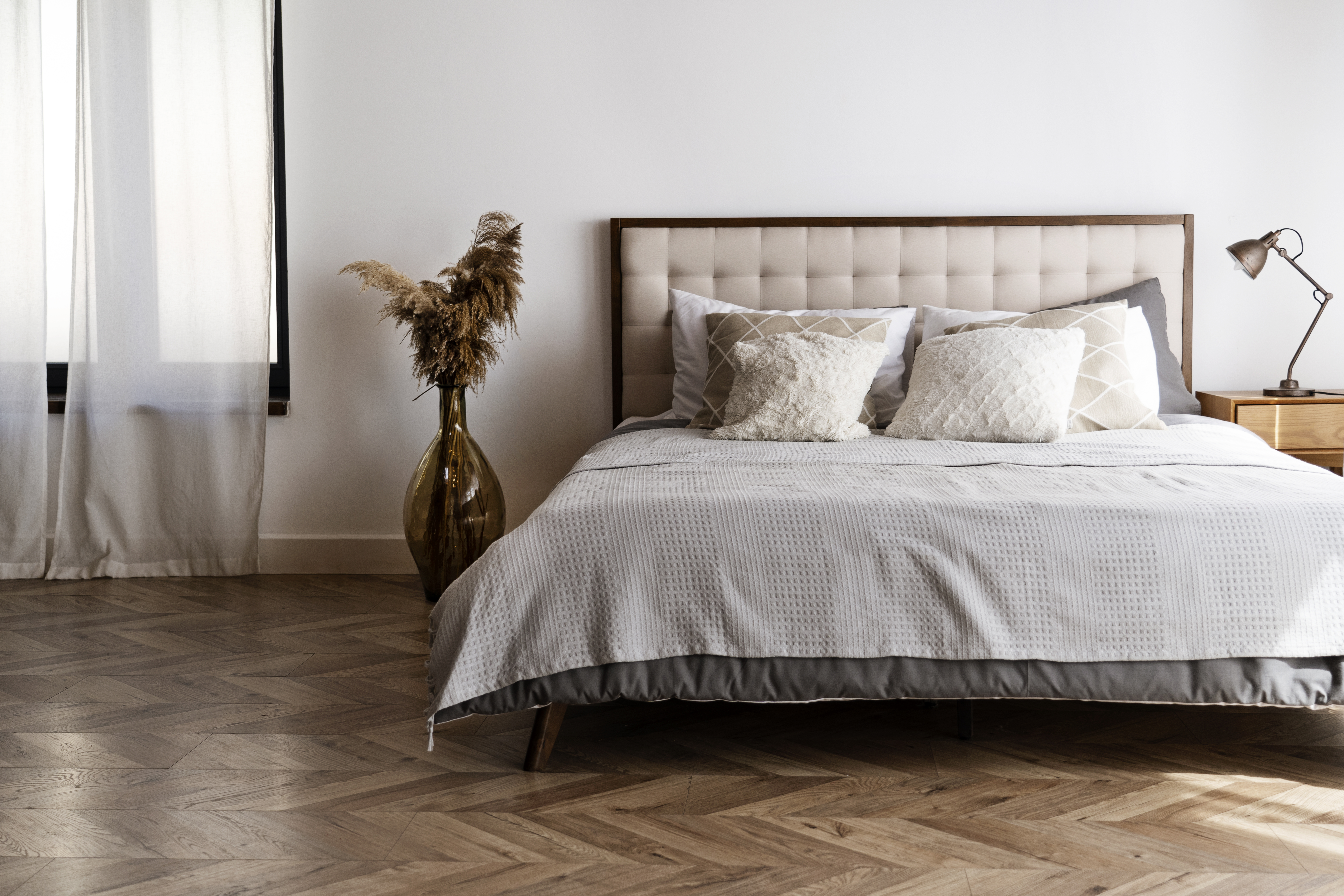 Quarto minimalista com cama de casal, cabeceira estofada bege, lençóis cinza e almofadas claras. À esquerda, um vaso alto de vidro âmbar com plantas secas e uma janela com cortina branca. À direita, uma mesa de cabeceira de madeira clara com um abajur de metal. O chão é de madeira com padrão espinha de peixe