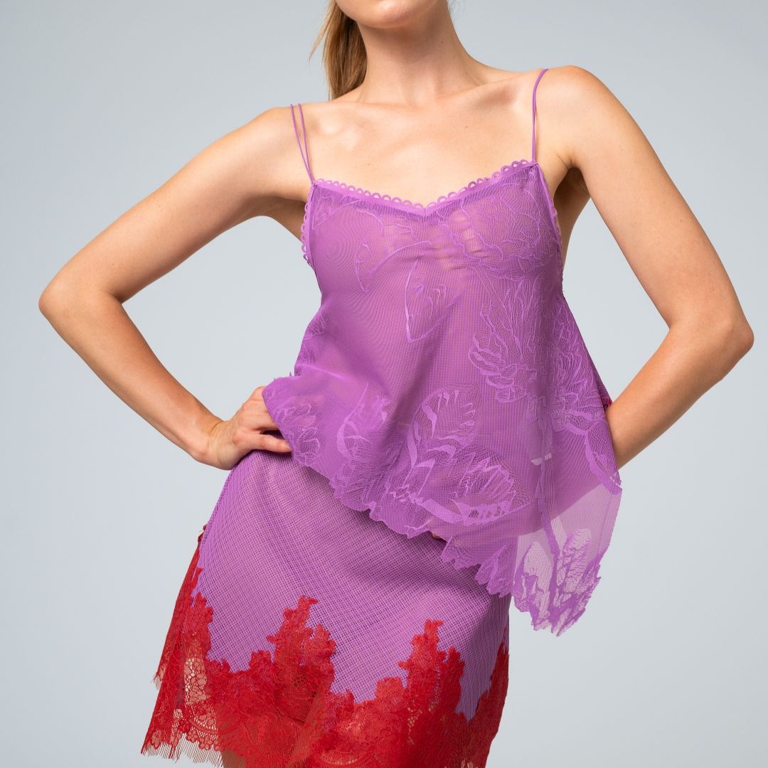 Mulher loira, com rabo de cavalo, posa com as m&atilde;os na cintura, vestindo um conjunto de lingerie roxa. A blusa de al&ccedil;as finas &eacute; transparente, com detalhes florais em renda, e a saia curta tem uma barra de renda vermelha vibrante.