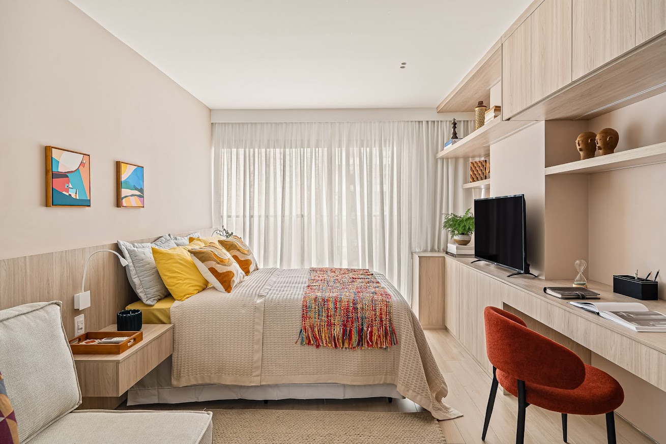 Quarto moderno com cama de casal, len&ccedil;&oacute;is claros, almofadas amarelas e uma manta colorida. &Agrave; esquerda, uma mesa de cabeceira com lumin&aacute;ria. &Agrave; direita, um painel de madeira clara com prateleiras, televis&atilde;o, e uma escrivaninha com cadeira laranja. Cortinas brancas cobrem a janela ao fundo