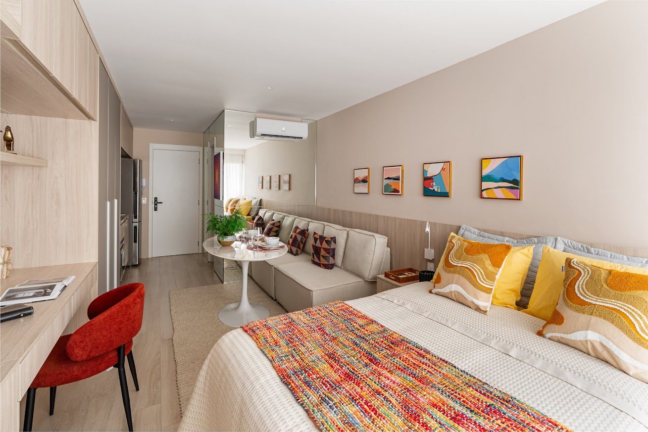 Est&uacute;dio moderno com cama de casal, manta colorida, almofadas amarelas, sof&aacute; bege com almofadas geom&eacute;tricas, mesa redonda com planta e lou&ccedil;a, e escrivaninha com cadeira vermelha. Arm&aacute;rios de madeira clara e parede bege com quadros abstratos completam o ambiente