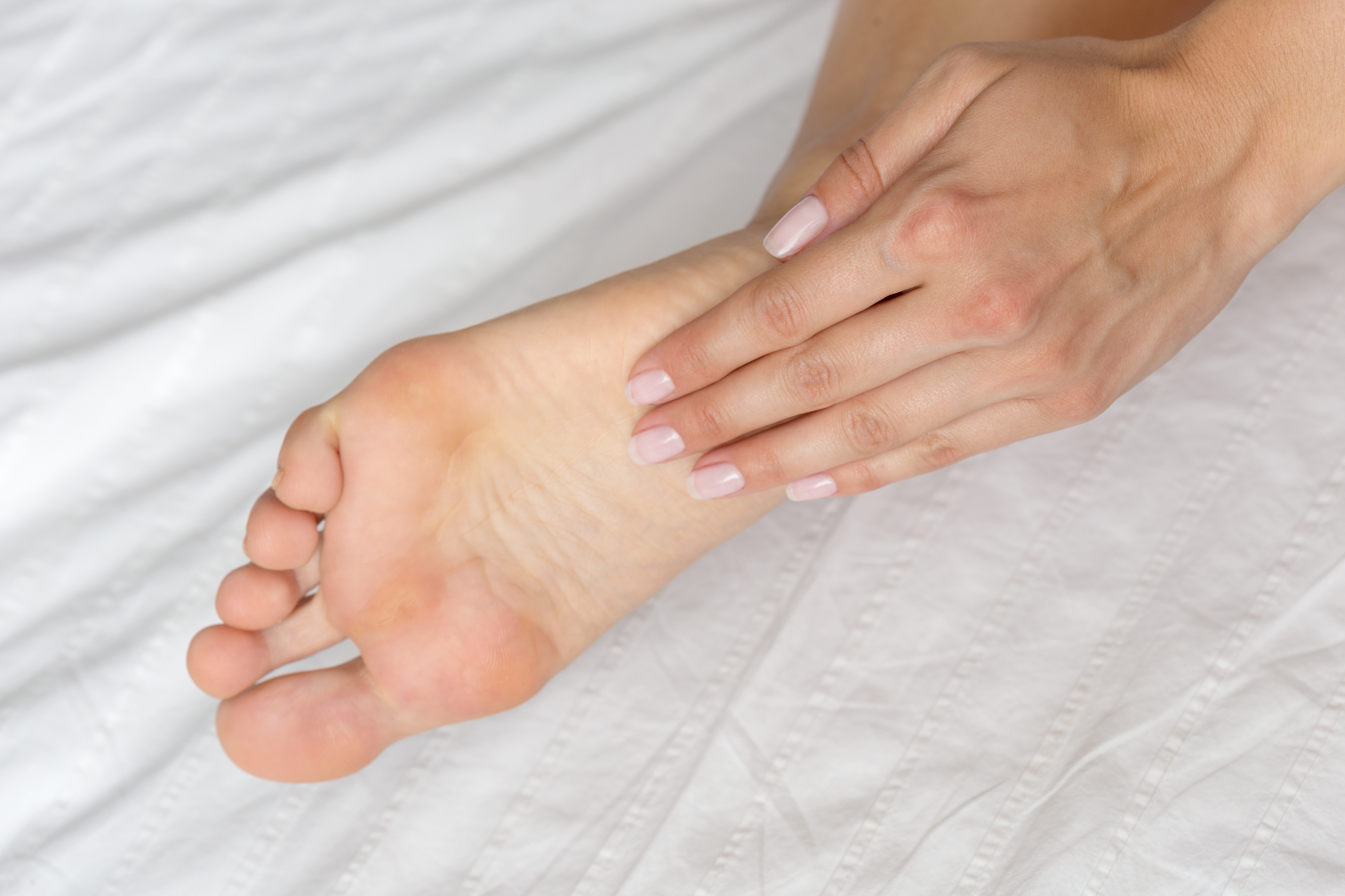 Uma mão feminina com unhas pintadas de rosa claro massageia a sola de um pé sobre um lençol branco