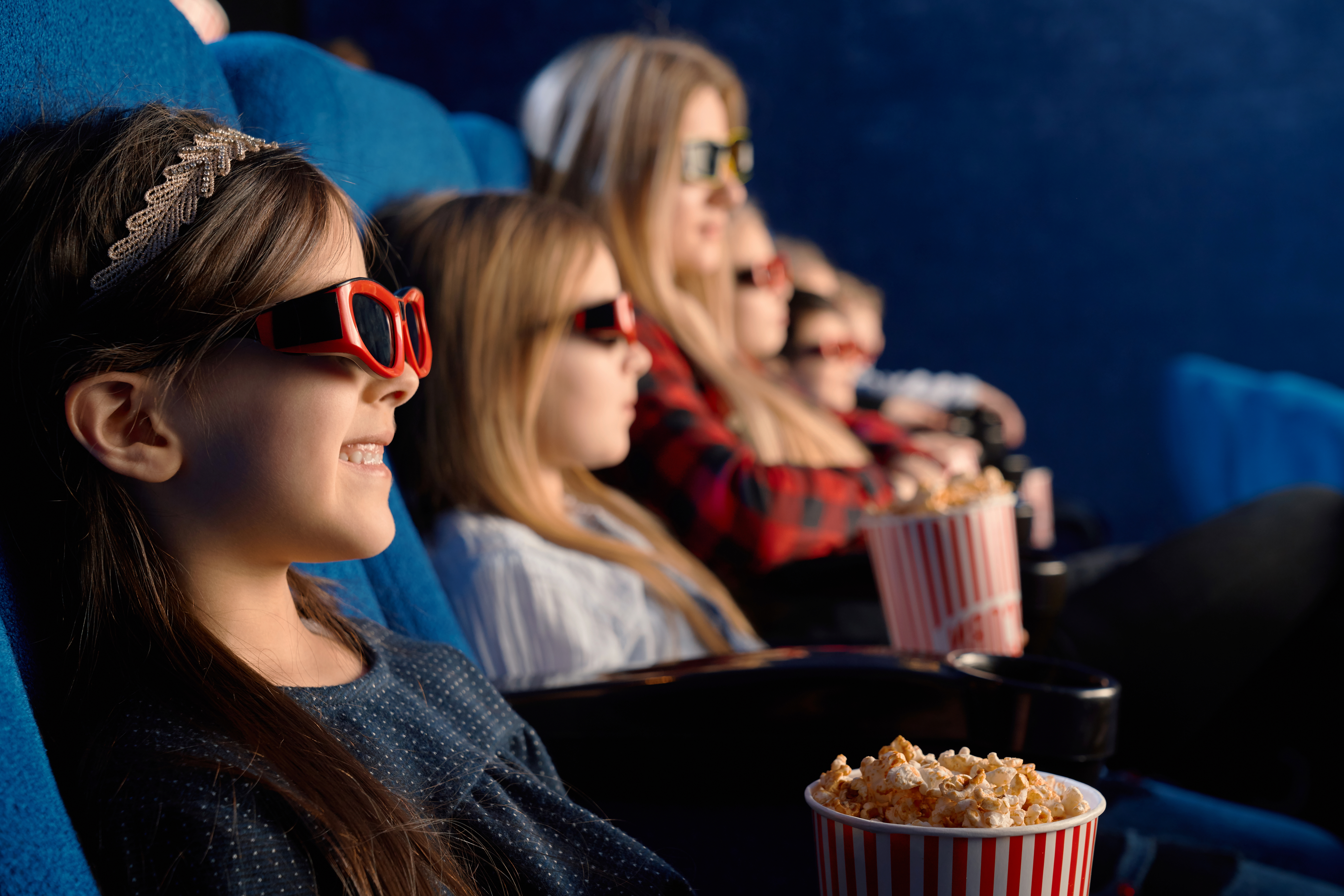 Crianças e adultos assistindo a um filme em 3D no cinema, usando óculos vermelhos e comendo pipoca em baldes listrados