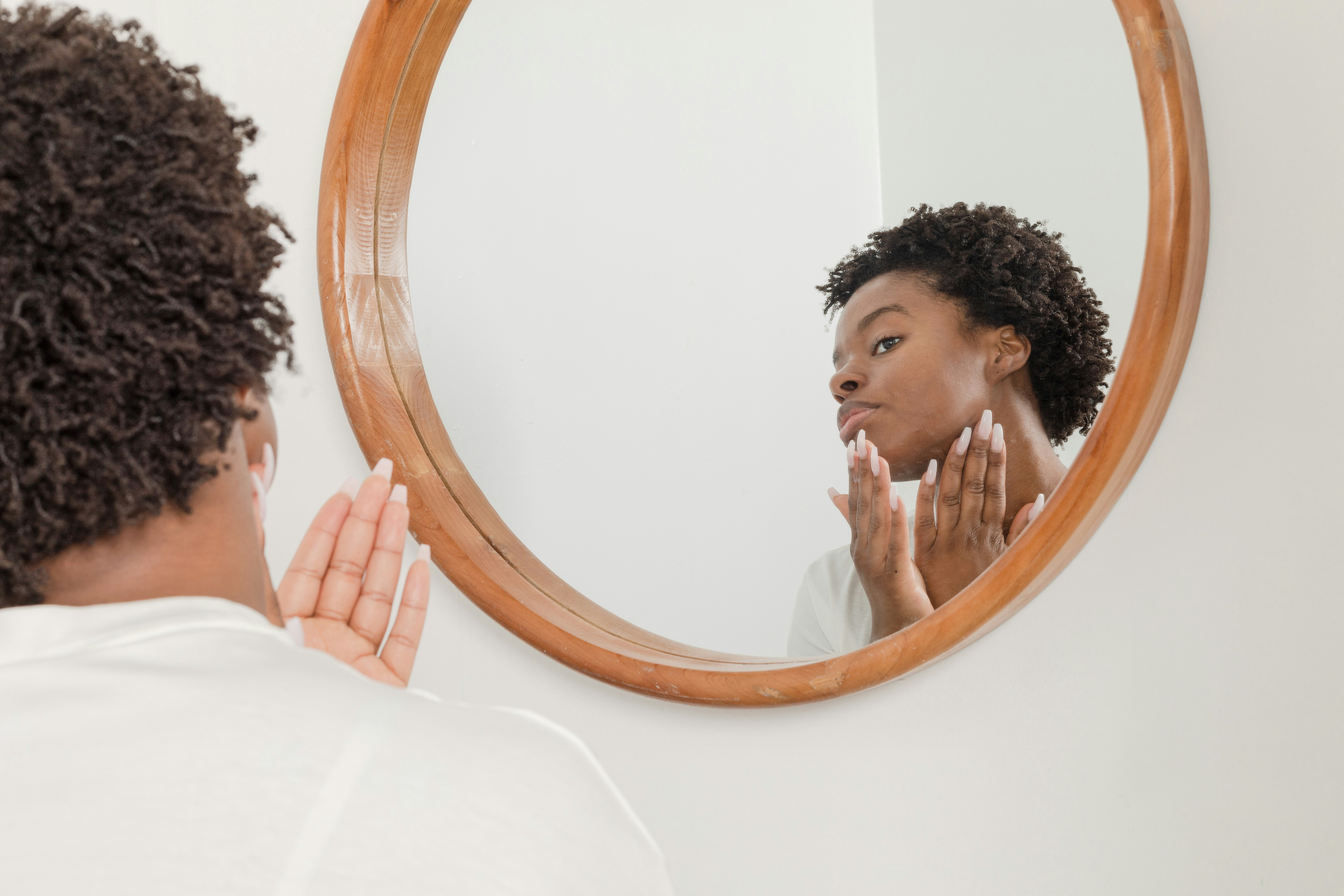 Mulher negra de cabelo crespo curto, de costas, olhando seu reflexo em um espelho redondo de madeira. Ela toca o rosto com as duas m&atilde;os, que t&ecirc;m unhas longas e claras, enquanto se observa no espelho. O fundo &eacute; branco