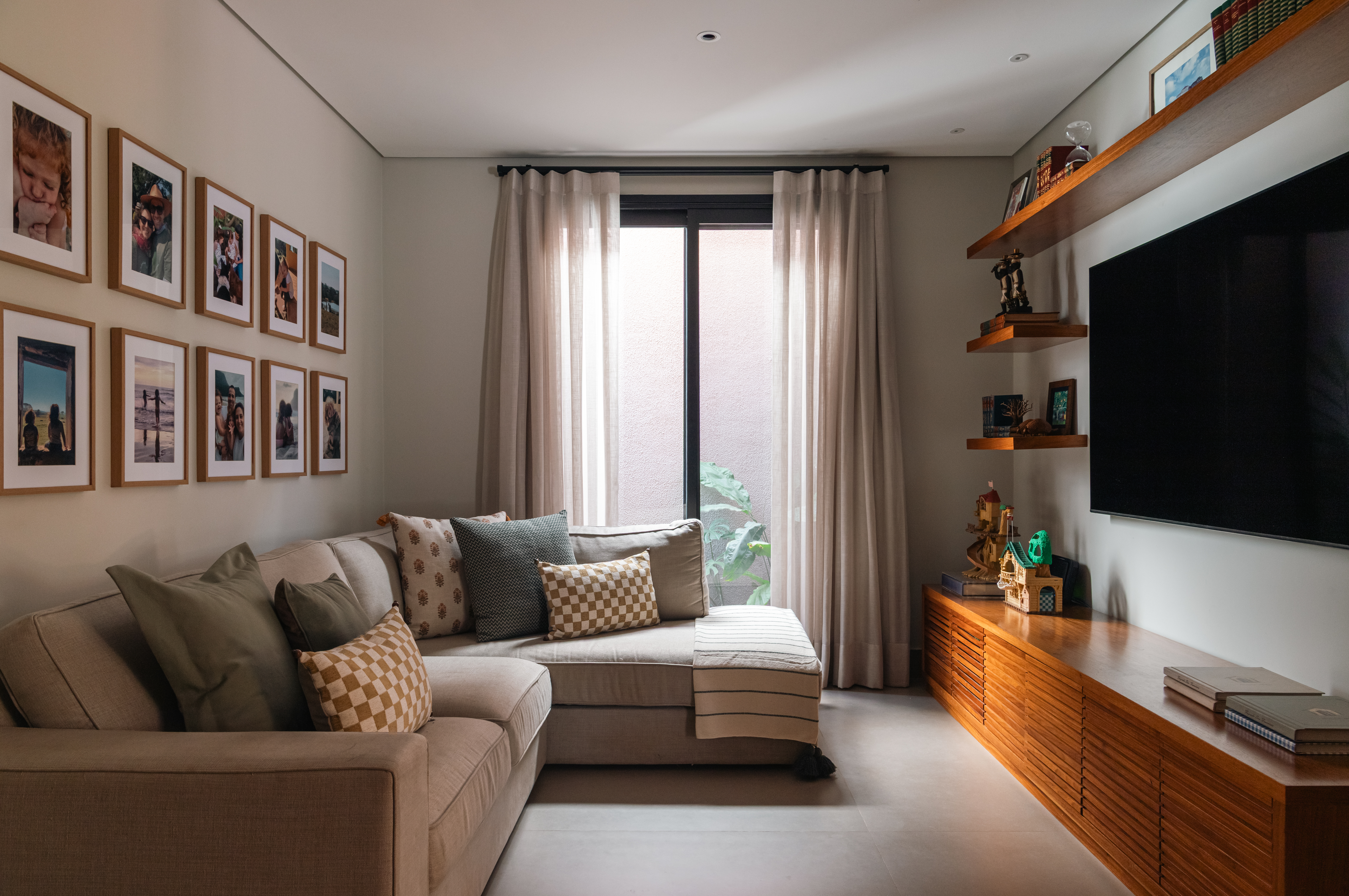 Sala de estar com sof&aacute; em L bege, almofadas variadas, e uma galeria de fotos emolduradas na parede. &Agrave; direita, uma TV preta sobre um rack de madeira com prateleiras flutuantes acima, decoradas com livros e objetos. Uma janela com cortinas claras ilumina o ambiente