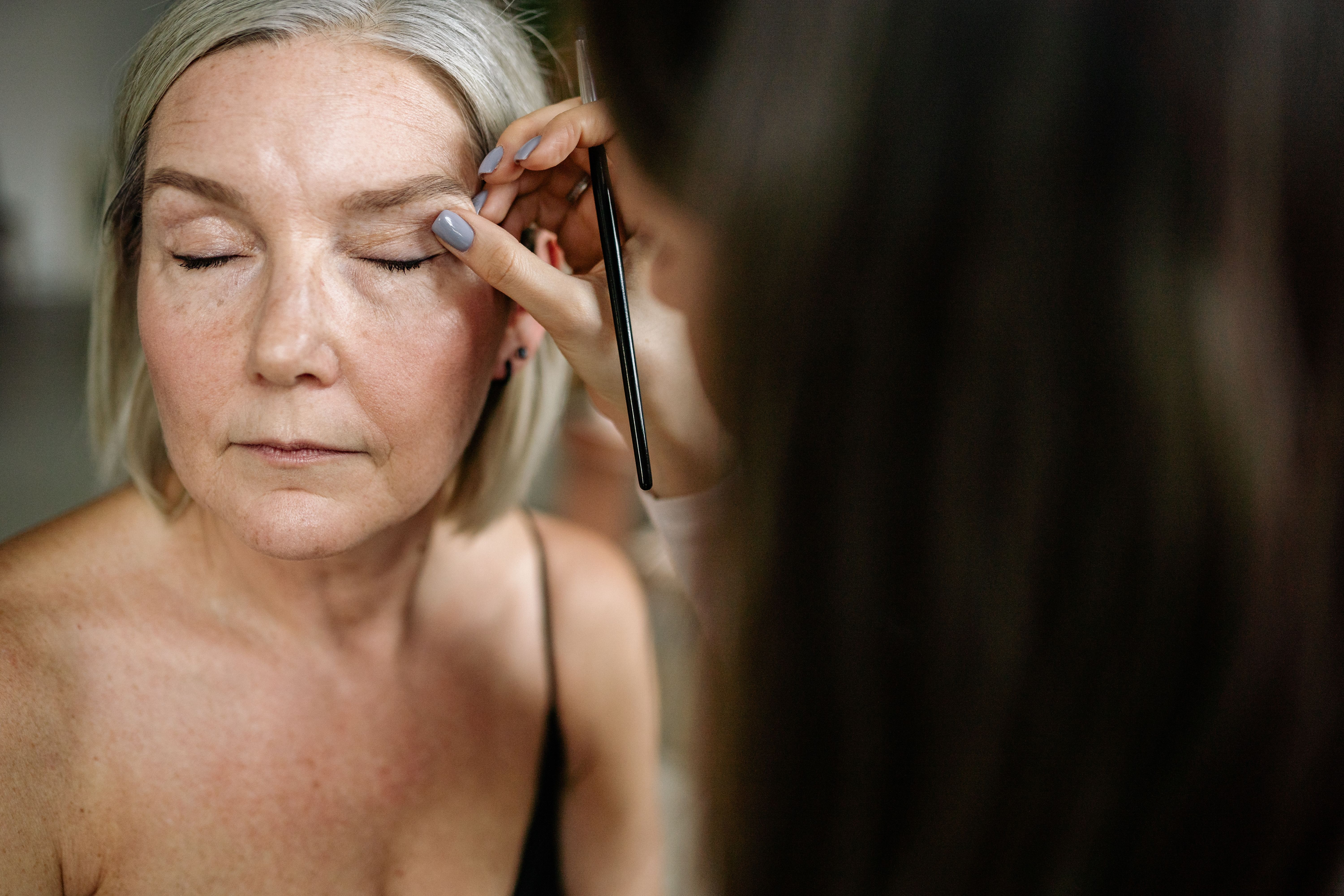 Mulher de cabelos grisalhos e olhos fechados tem a sobrancelha maquiada por uma mão com unhas cinzas segurando um pincel fino