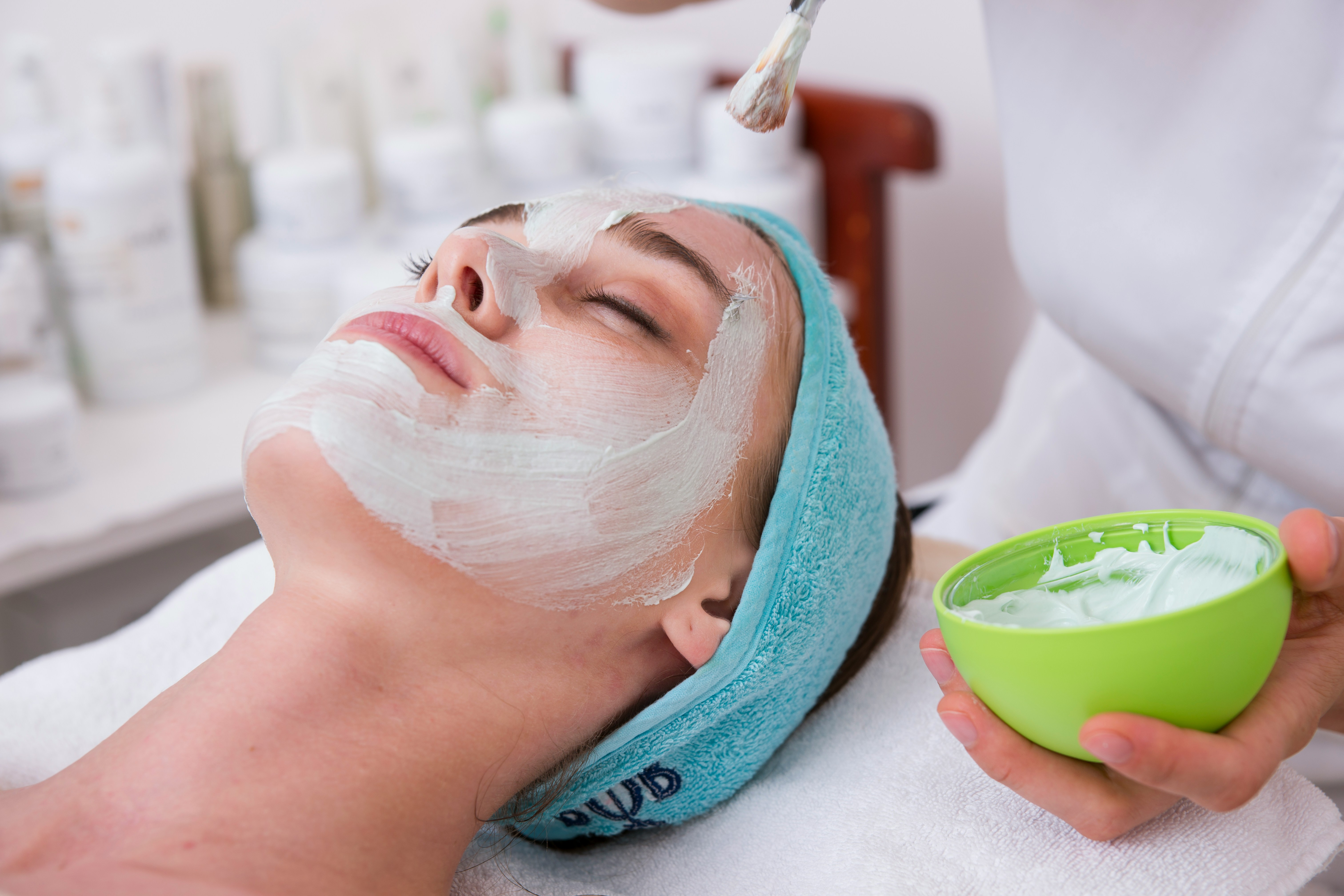 Mulher deitada com os olhos fechados, recebendo uma m&aacute;scara facial branca. Ela usa uma faixa de cabelo azul e uma esteticista aplica o produto com um pincel, segurando uma tigela verde com mais creme