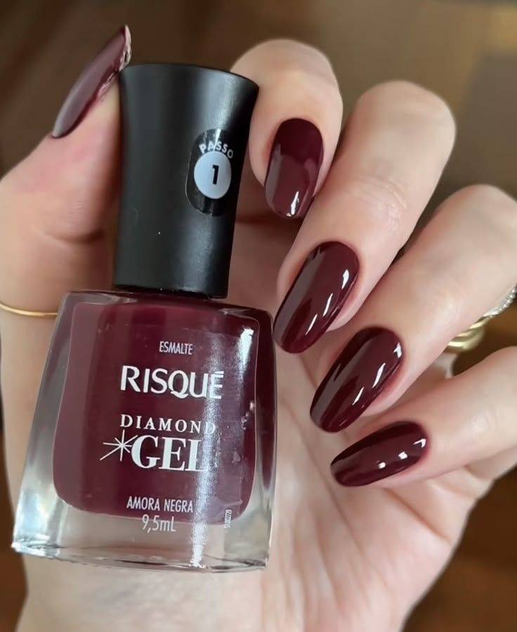 M&atilde;o feminina com unhas pintadas de esmalte vinho escuro, segurando um frasco de esmalte Risqu&eacute; Diamond Gel Amora Negra