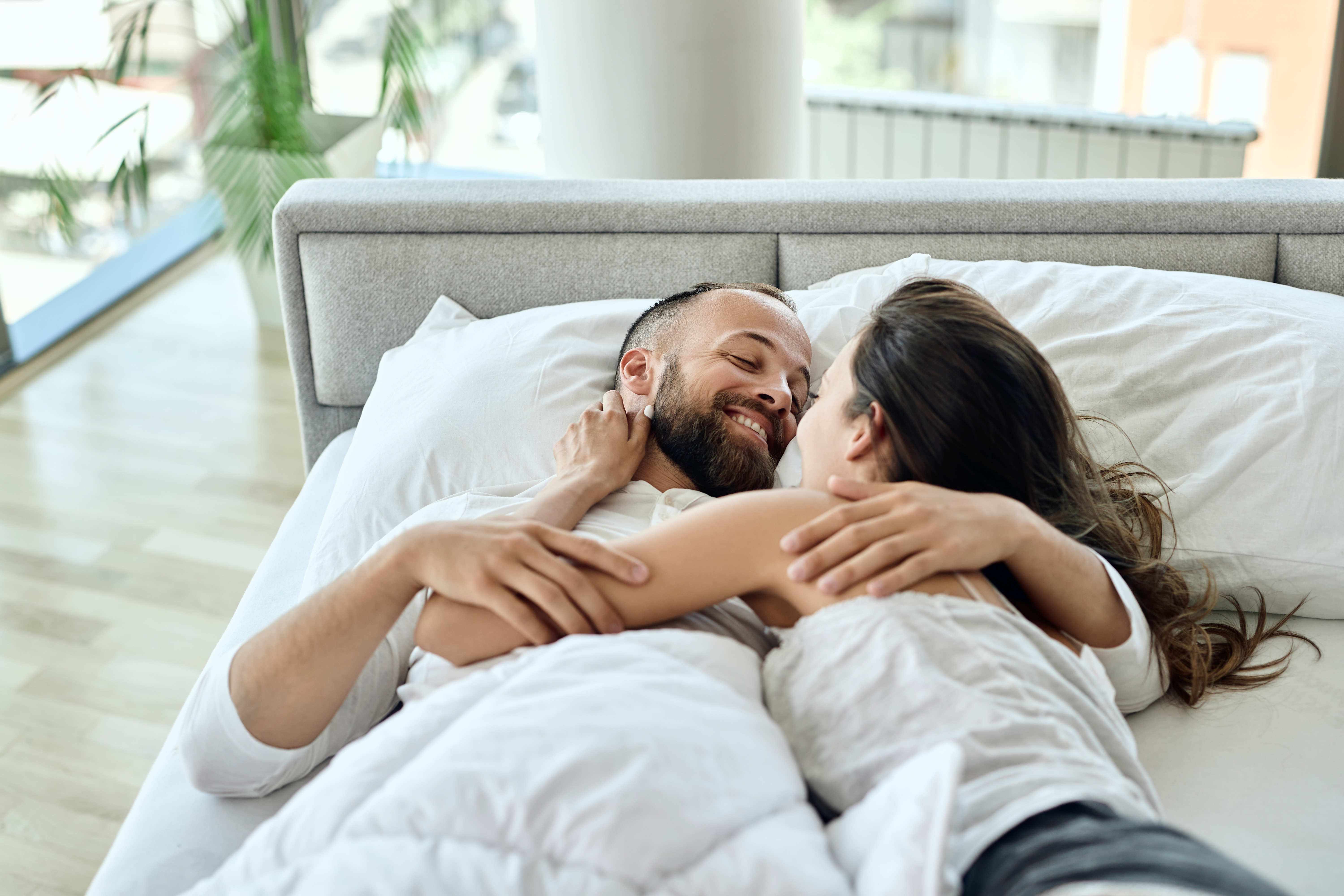 Casal sorridente e abra&ccedil;ado na cama, com o homem barbudo de olhos fechados e a mulher com cabelo escuro, ambos sob len&ccedil;&oacute;is brancos. O quarto tem uma janela grande e piso de madeira ao fundo