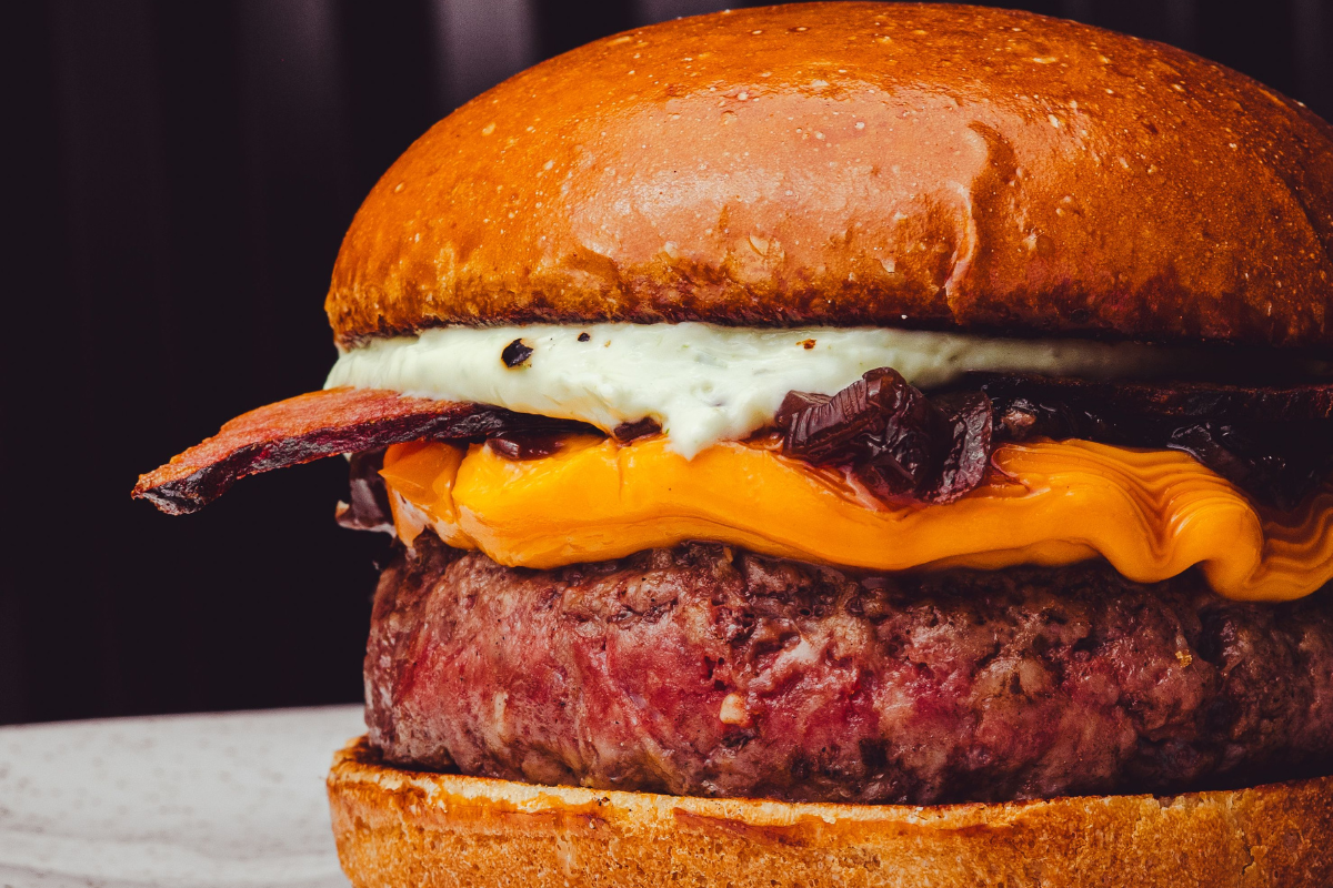 Hambúrguer gourmet com pão brioche tostado, queijo cheddar derretido, bacon crocante, cebola caramelizada e molho branco cremoso, em fundo escuro