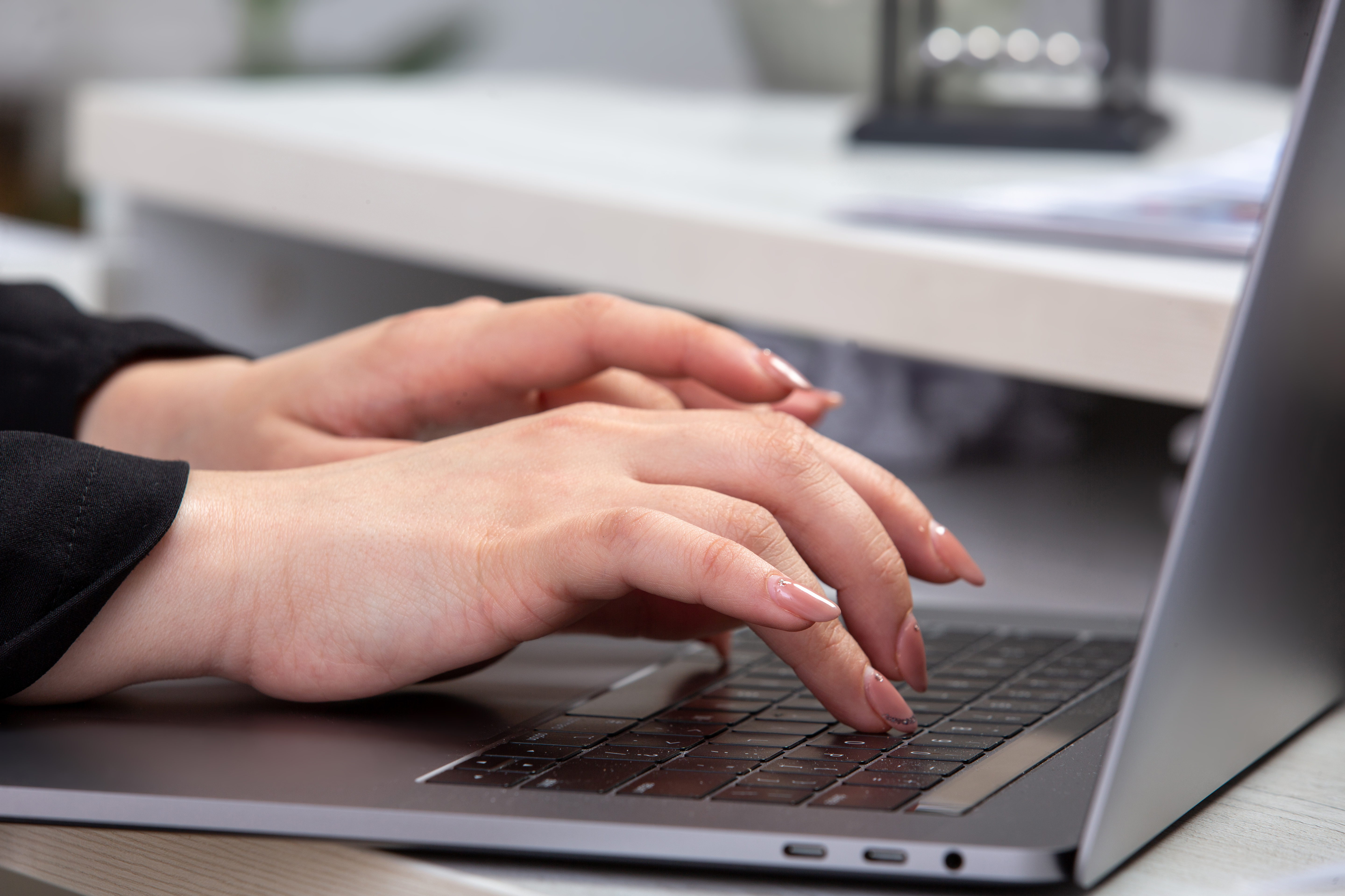 M&atilde;os femininas com unhas alongadas digitando em um teclado de notebook cinza-escuro, com a tela do computador parcialmente vis&iacute;vel &agrave; direita e um fundo de escrit&oacute;rio desfocado